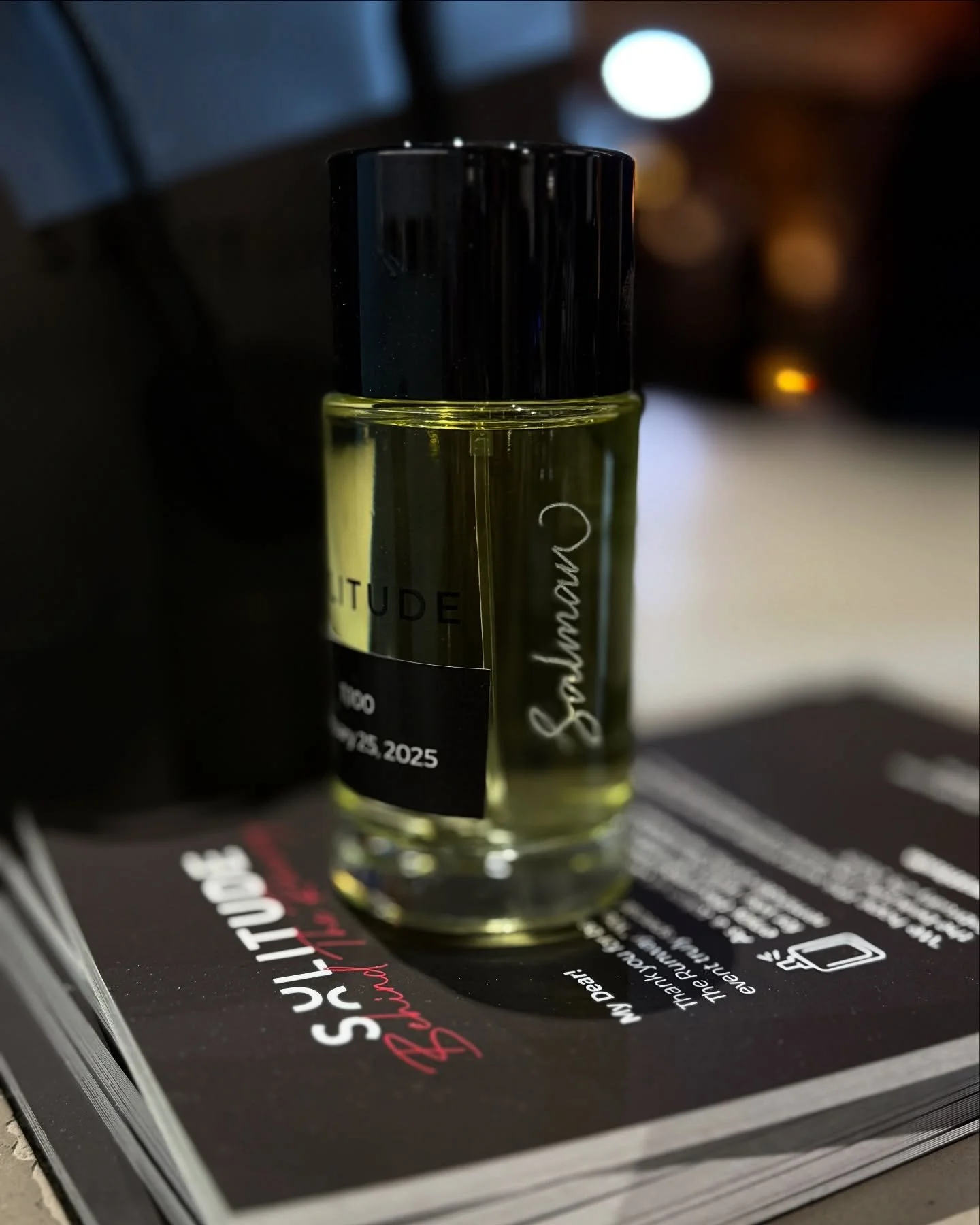 A small spray bottle of perfume or essential oil with a black cap is placed on top of a magazine or booklet with text and images. The bottle has a yellowish liquid inside.