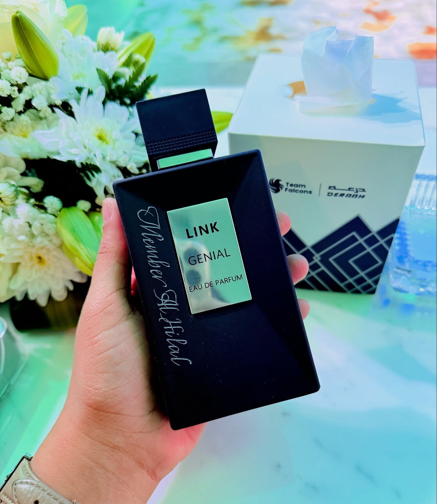 A hand holding a black box of perfume labeled 'LINK GENIAL EAU DE PARFUM' with a floral bouquet and a white gift box with tissue paper inside in the background.