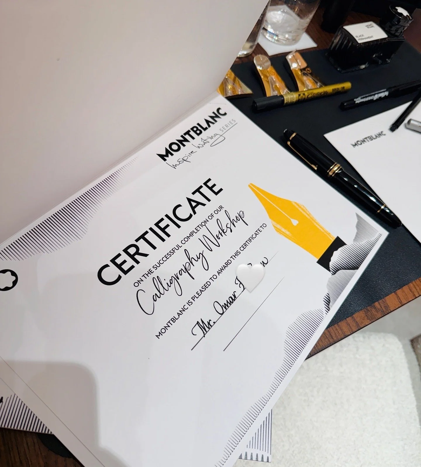 A Montblanc certificate for completing Calligraphy Workshop, placed on a table with pens, markers, a glass of water, and office supplies.