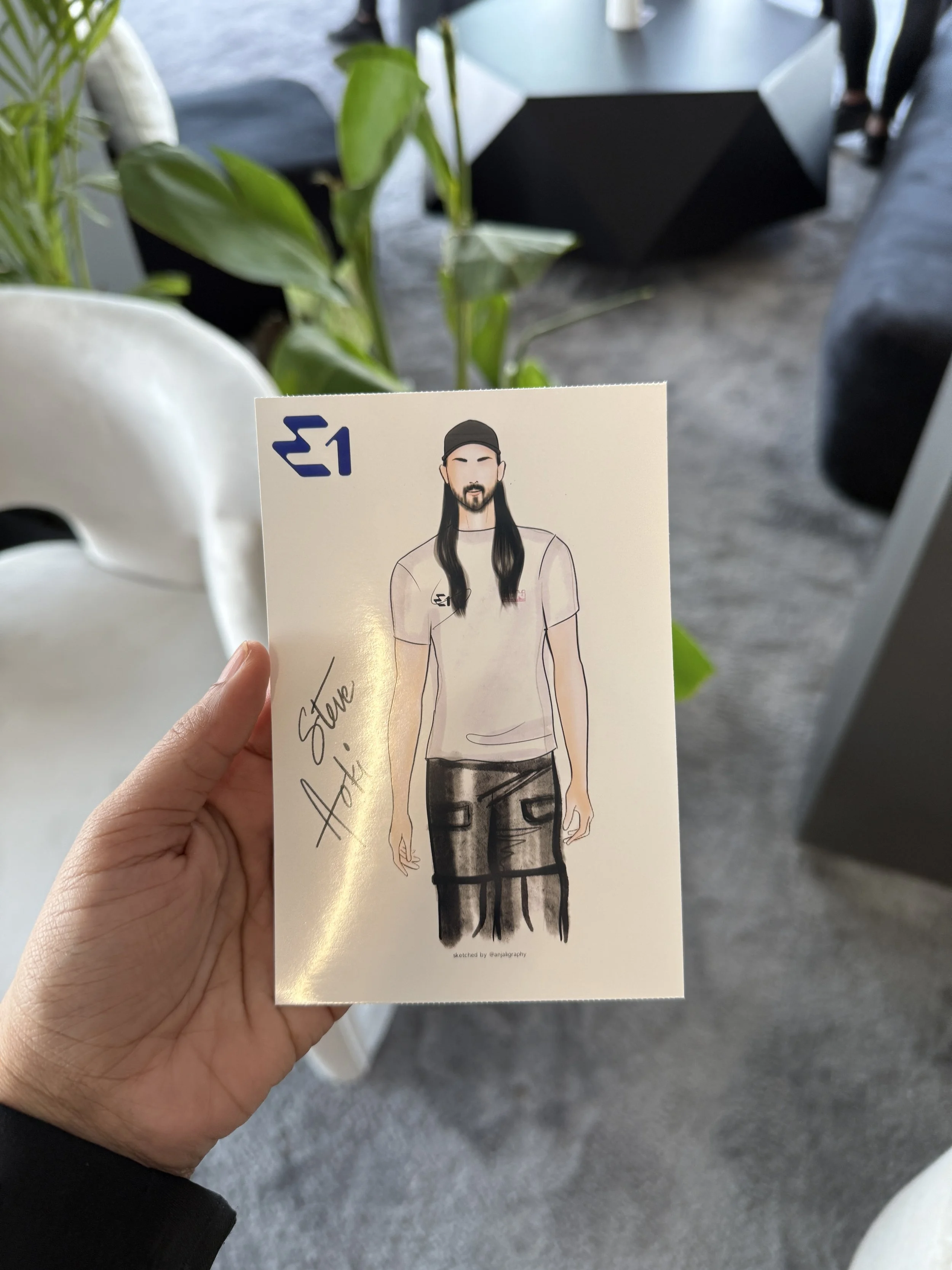 Hand holding a printed illustration of a man with long dark hair, a beard, wearing a black cap, a light-colored t-shirt, and dark pants, in an indoor setting with plants and modern furniture.