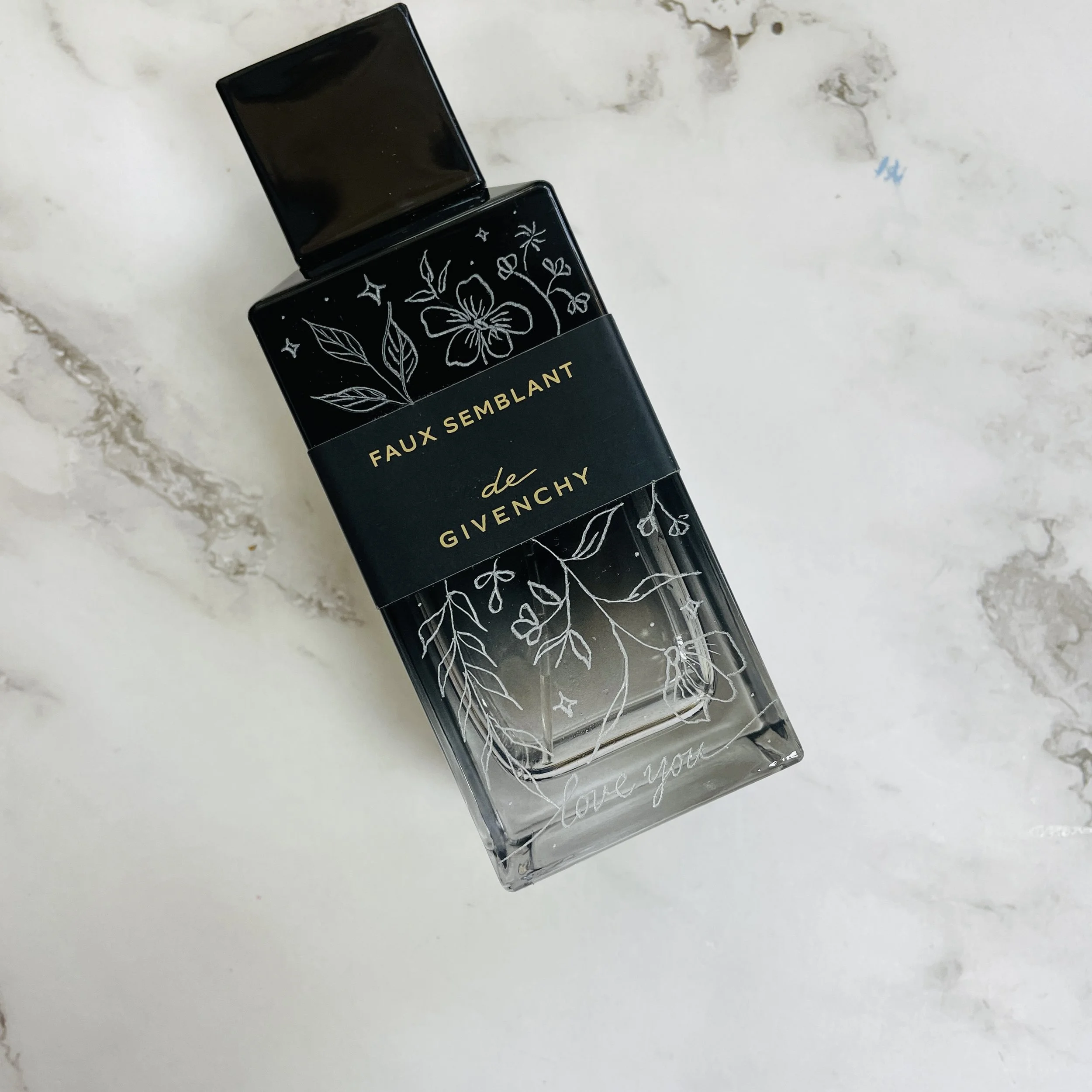A black perfume bottle with a floral and star design on it, placed on a white marble surface.