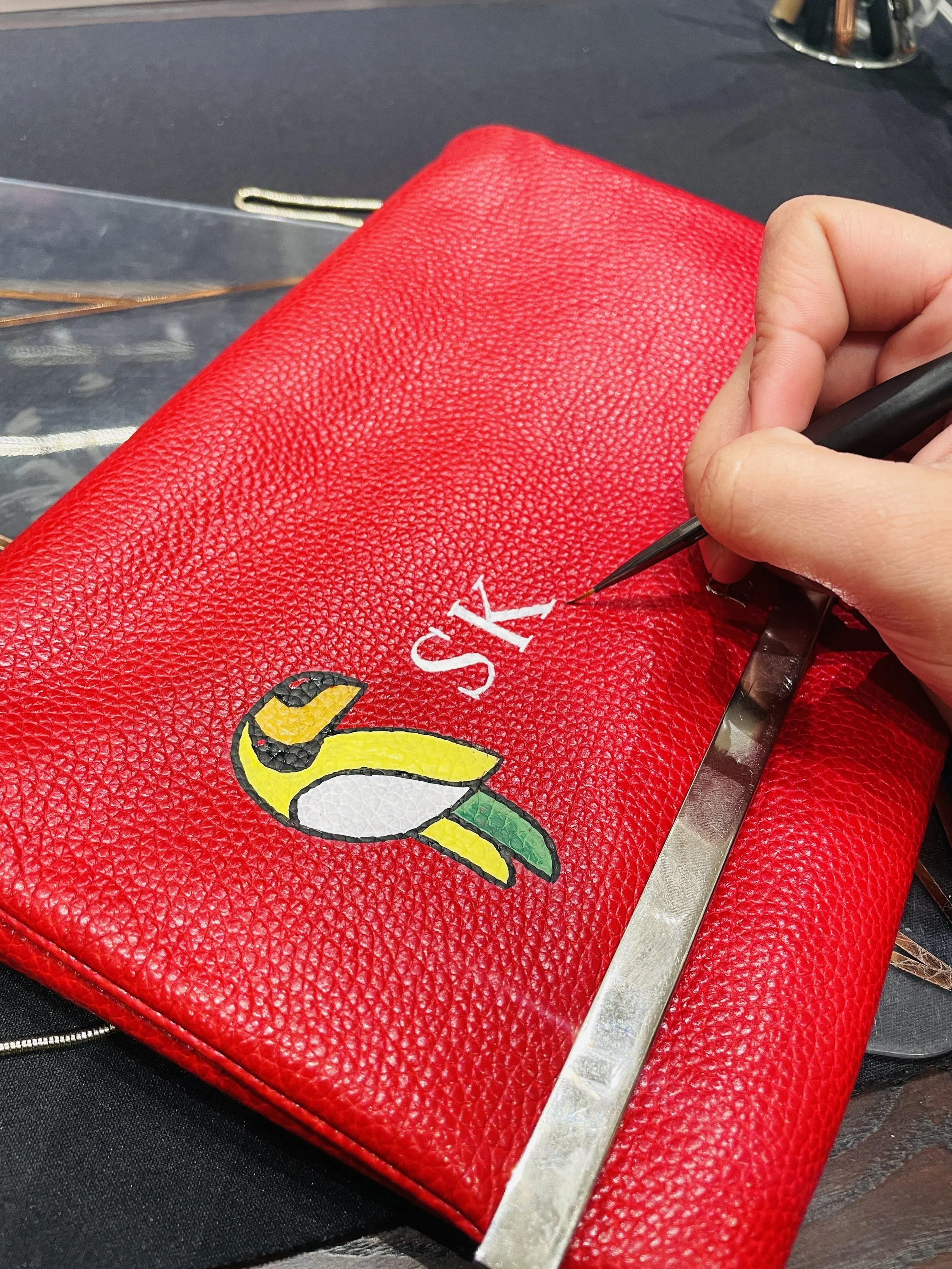 A person's hand holding a black pen, writing on a red leather bag with a cartoon character and white text. The bag is resting on a black surface.