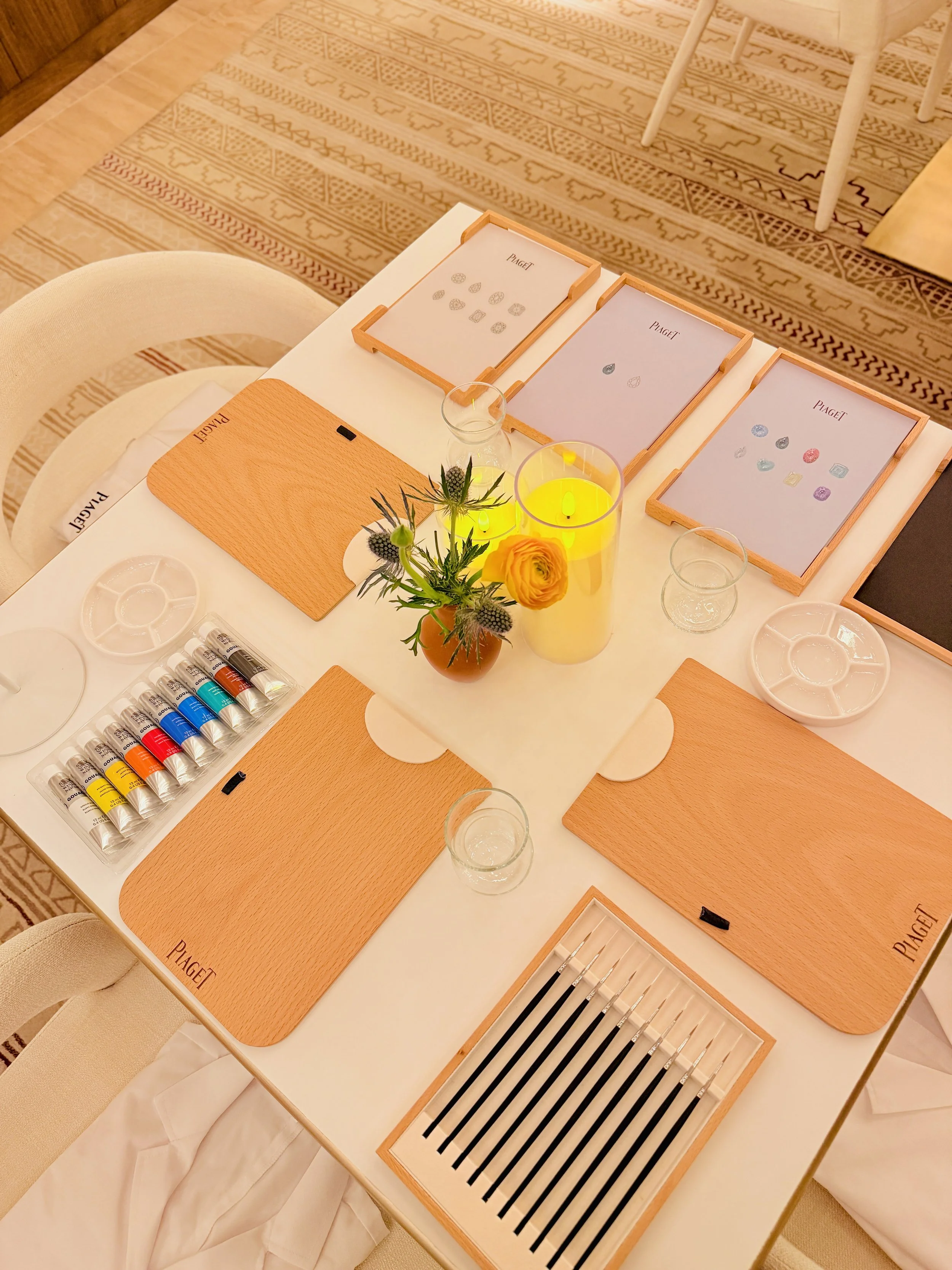Table setup for a jewelry or gemstone testing event with wooden trays, paint tubes, and a small flower arrangement with yellow and purple flowers in a yellow vase.