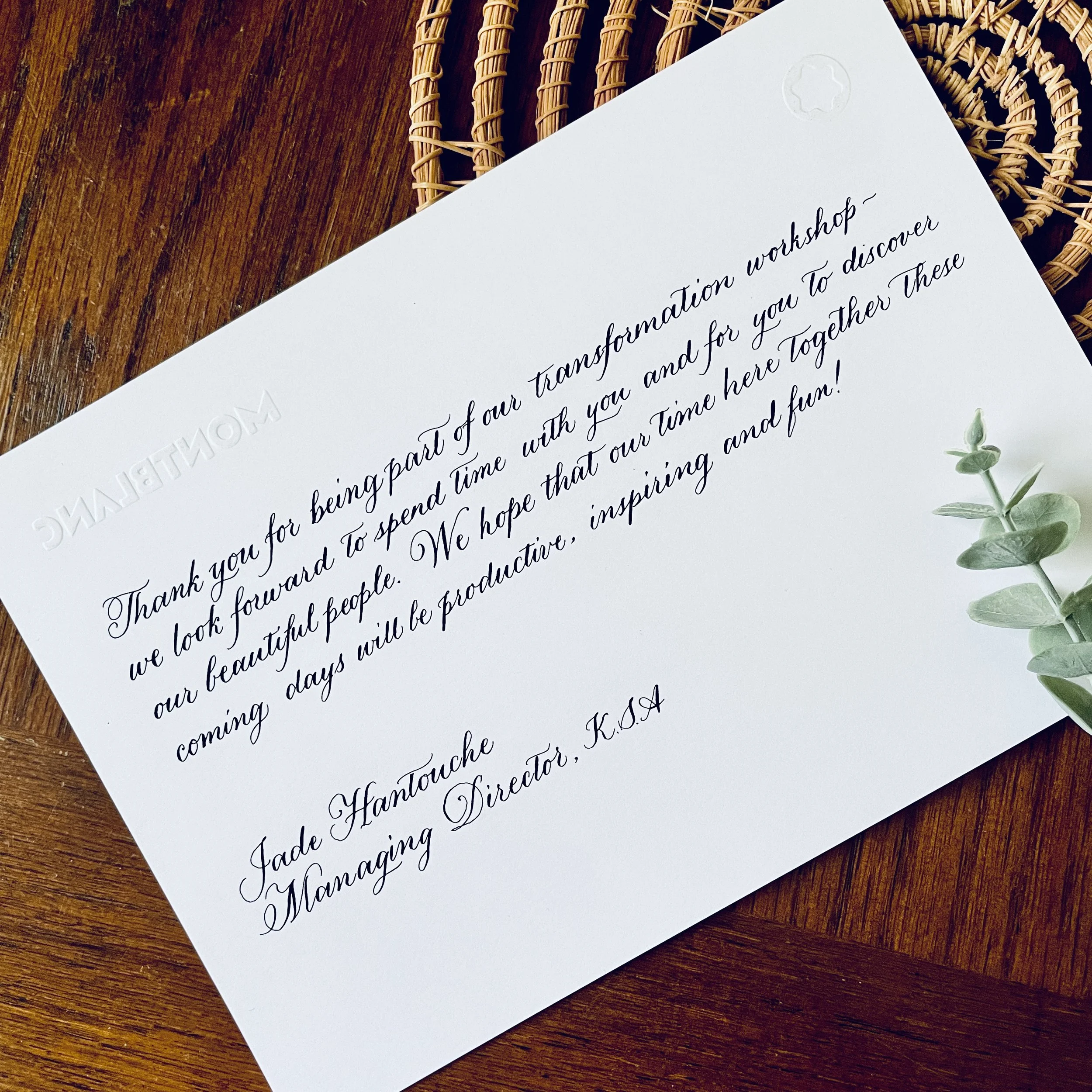 Close-up of a white letter with elegant cursive writing, placed on a wooden surface, with a small sprig of greenery beside it and a wicker placemat underneath.