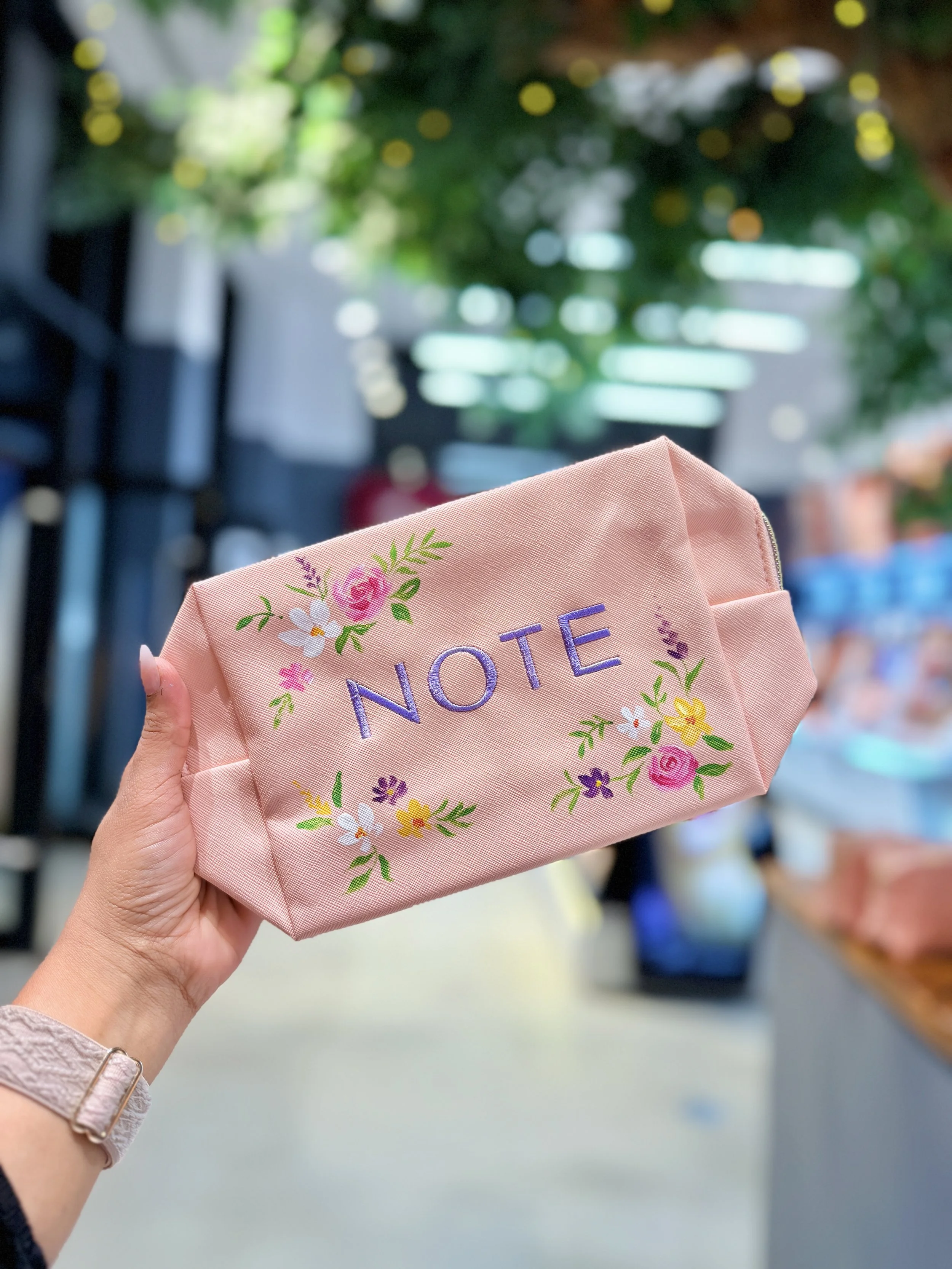 A hand holding a pink cloth face mask with the word 'NOTE' embroidered in purple, surrounded by colorful floral embroidery, in a store aisle with blurred products in the background.