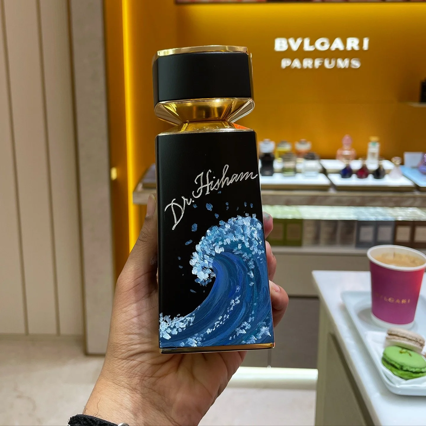 A person holding a black bottle with a blue wave design and the words "Dr. Fisham" written on it, in a store with a "Bvlgari Perfums" sign in the background. There are small bottles on display and a tray with macarons and a cup of coffee.