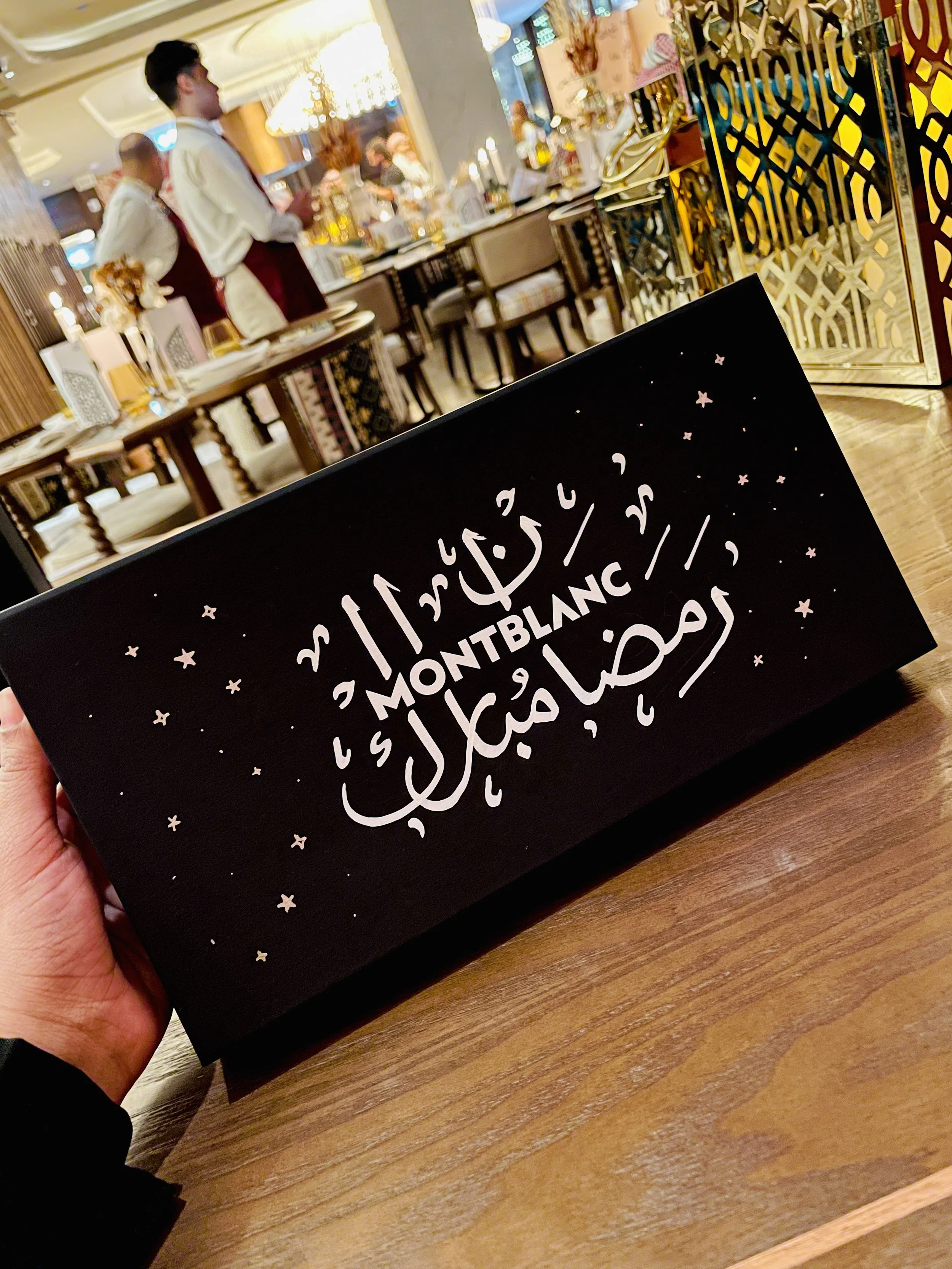 A person holding a black card with white Arabic and English text that reads 'MONTBLANC' and 'Ramadan Mubarak,' with small stars and crescent moon decorations, in a restaurant with waitstaff and diners in the background.