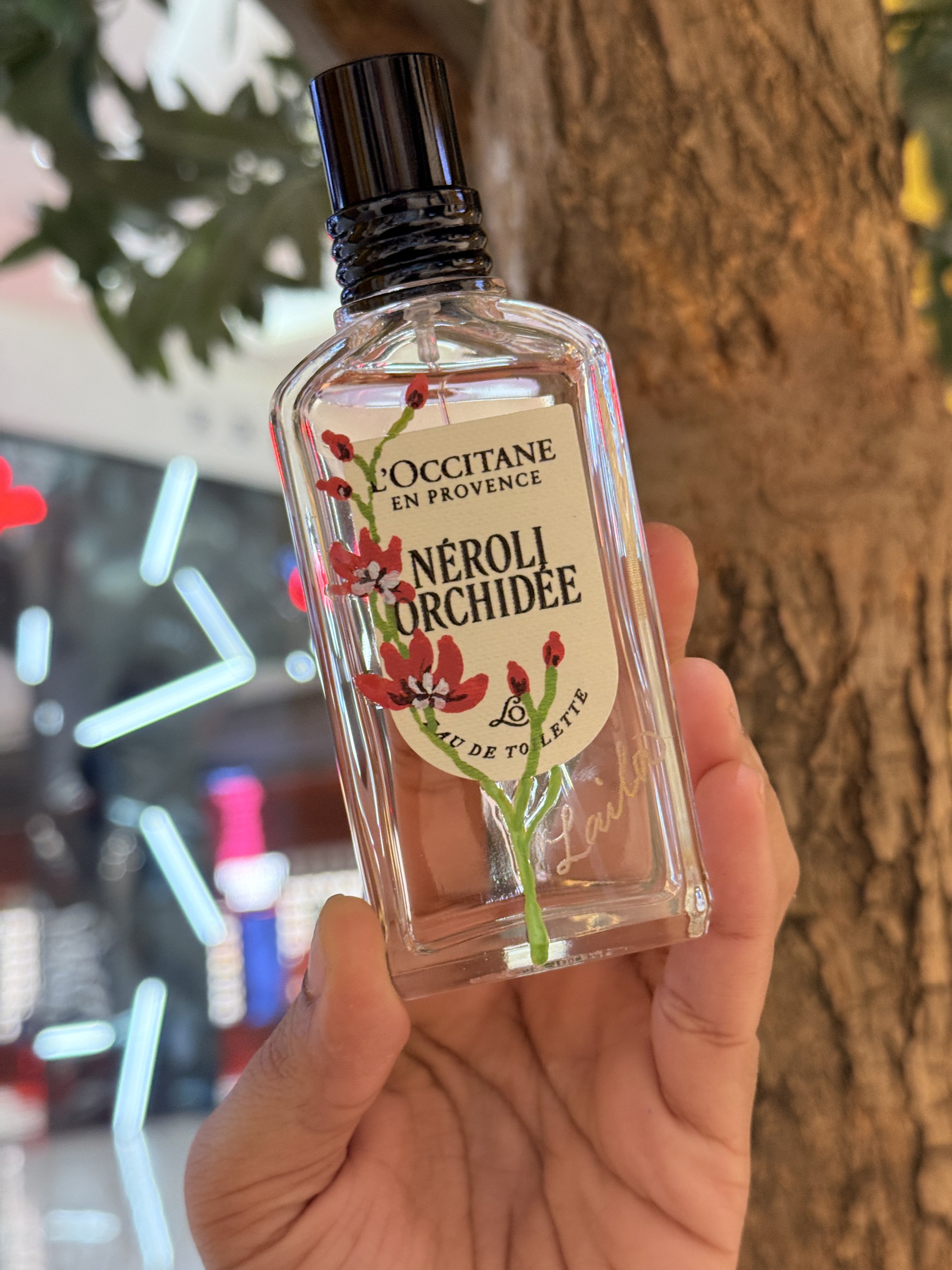 A person holding a clear perfume bottle labeled 'L'OCCITANE EN PROVENCE NÉROLI ORCHIDÉE' with a floral design, standing in front of a tree trunk with blurred colorful lights in the background.
