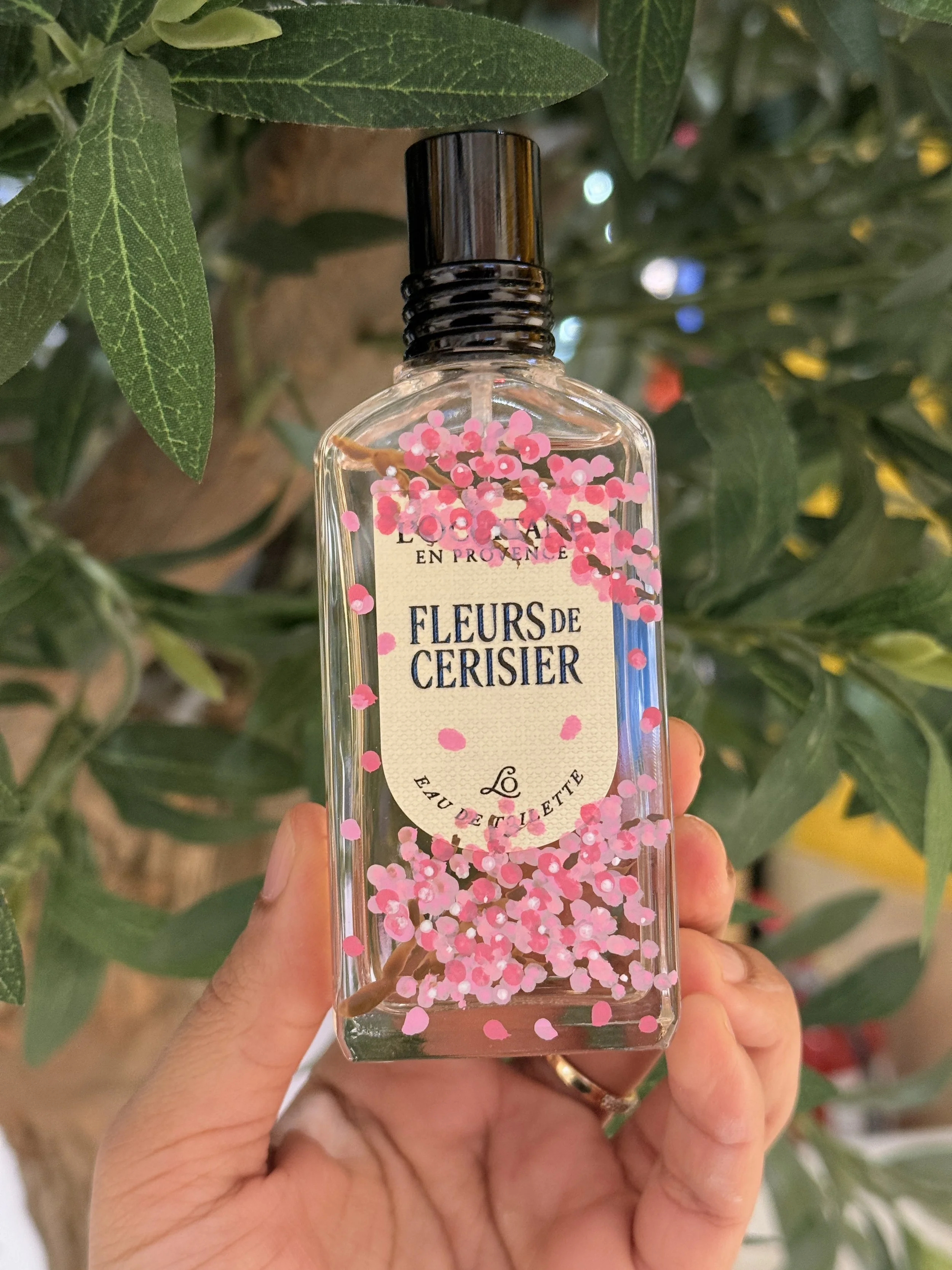 A person's hand holding a clear glass spray bottle labeled 'Fleurs de Cerisier' (Cherry Blossom) with pink cherry blossom petals floating inside the bottle. Green leaves and branches are in the background.