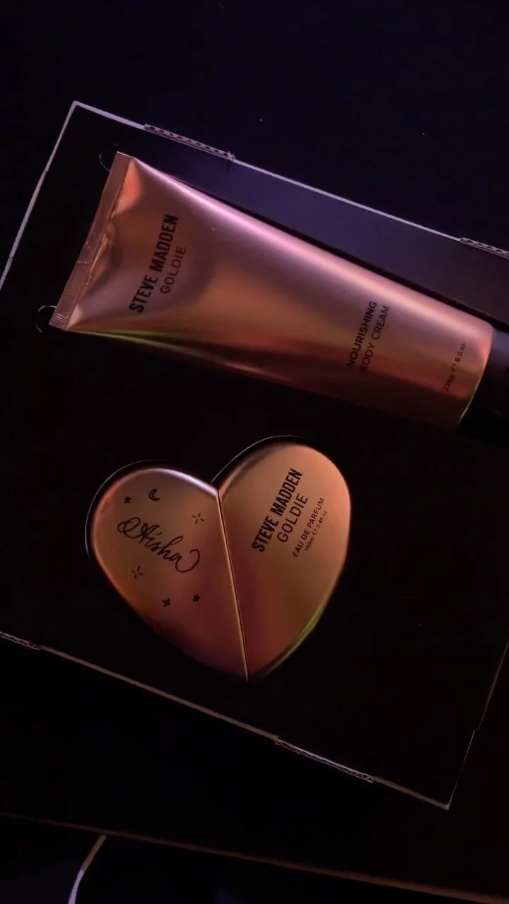 A gift set containing a tube of Steve Madden Goldie nourishing body cream and a heart-shaped perfume called "Arisha," both in a black Gift box.