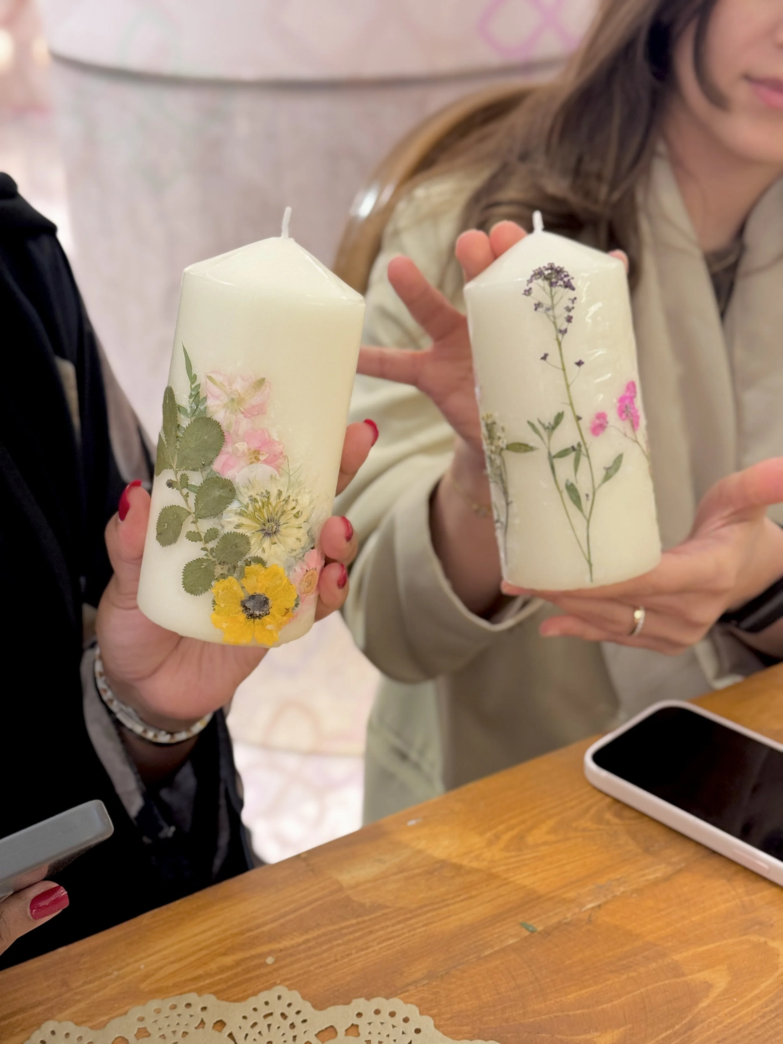 Two women holding white pillar candles decorated with pressed flowers, seated at a wooden table with a smartphone and a paper doily, indoors with blurred background.