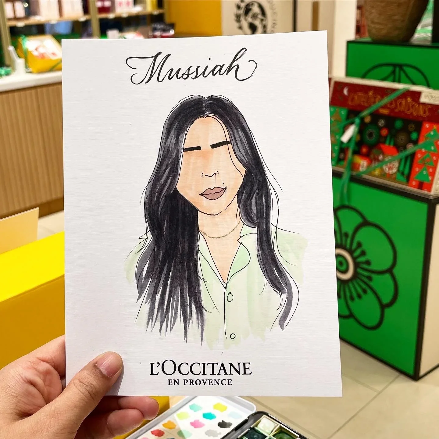 Hand holding an illustrated portrait of a woman with long black hair, wearing a light-colored shirt, with the text 'Mussiah' at the top and 'L'OCCITANE EN PROVENCE' at the bottom, in a store with shelves of colorful products and a green flower design