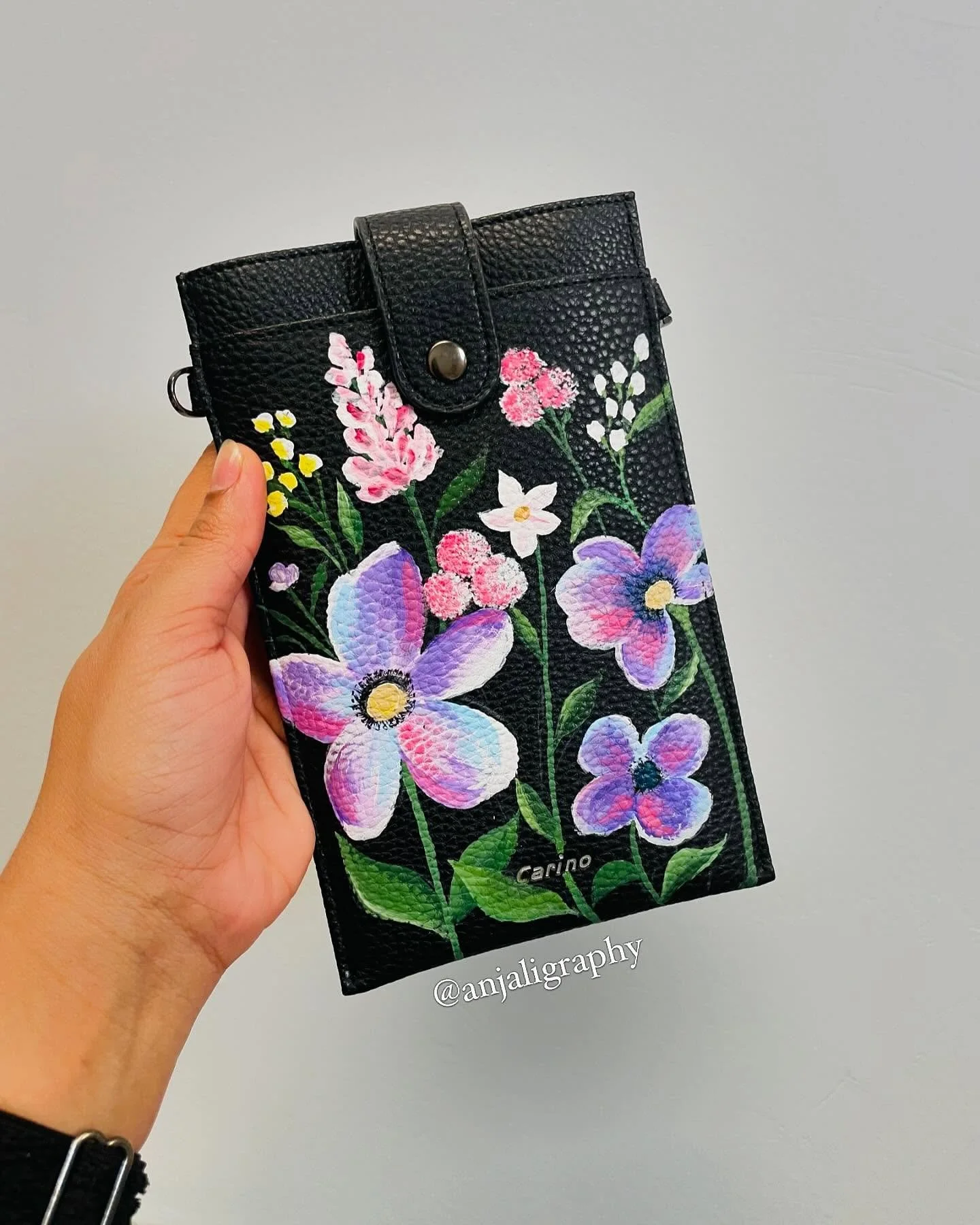 A person's hand holding a black leather wallet decorated with colorful painted flowers on a plain light gray background.