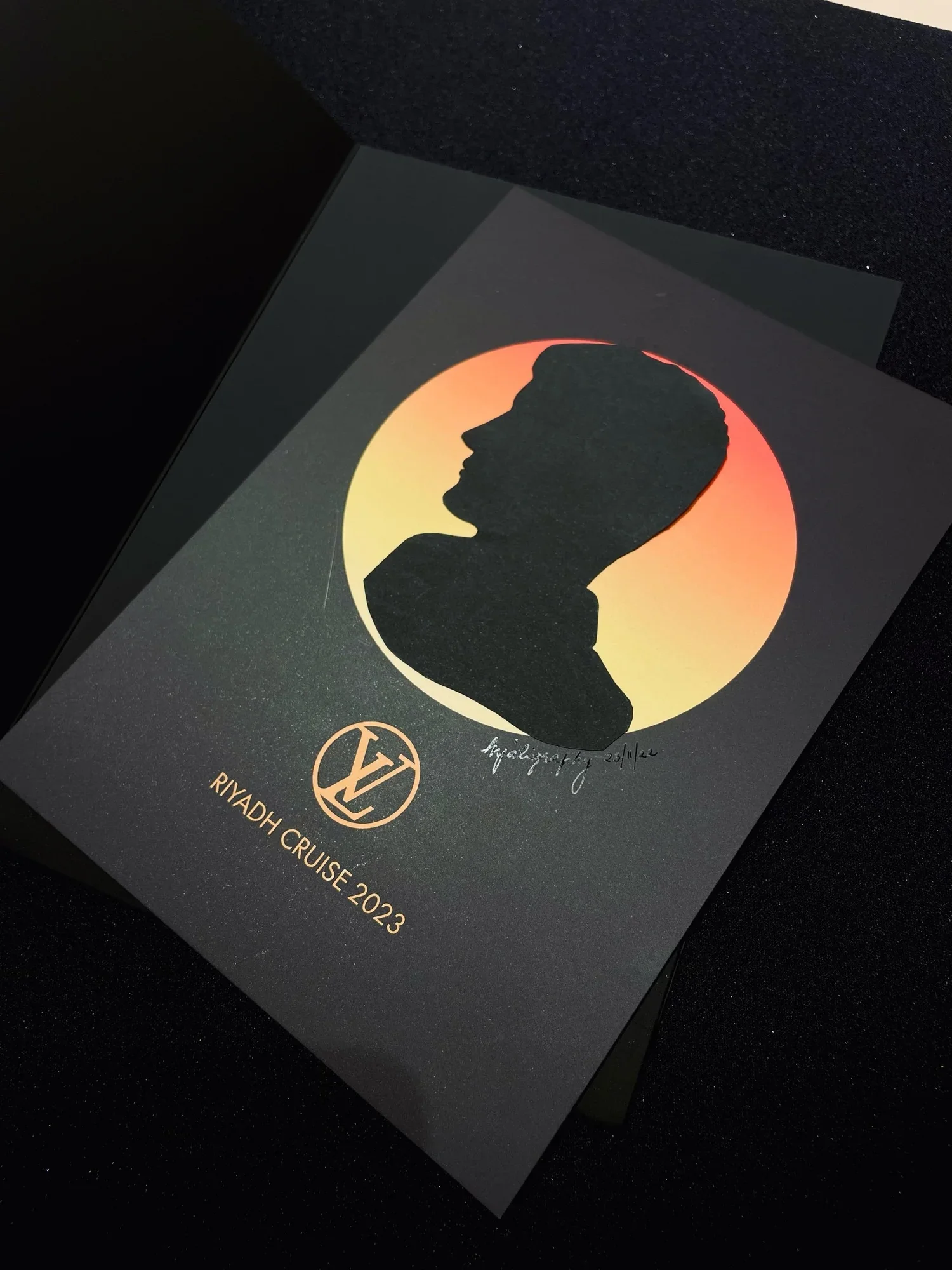 A black brochure or card with a silhouette of a person's profile inside a sunset-colored circle, text including 'RYADH CRUISE 2023' and a signature with the date 25/4/20.