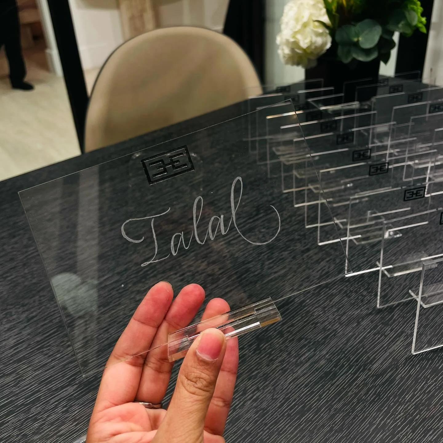 Hand holding a clear acrylic sign with the name 'Jala' written in cursive, placed on a black table near a chair and a vase of flowers.