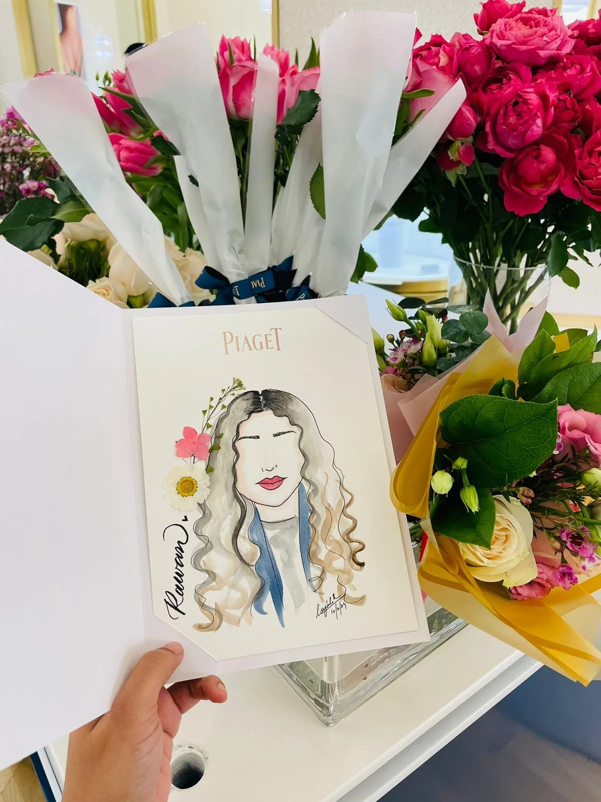 A hand holding a card with a caricature of a woman with long wavy hair, red lips, and a blue scarf, surrounded by flowers. Behind are bouquets of pink, white, and purple flowers on a table.