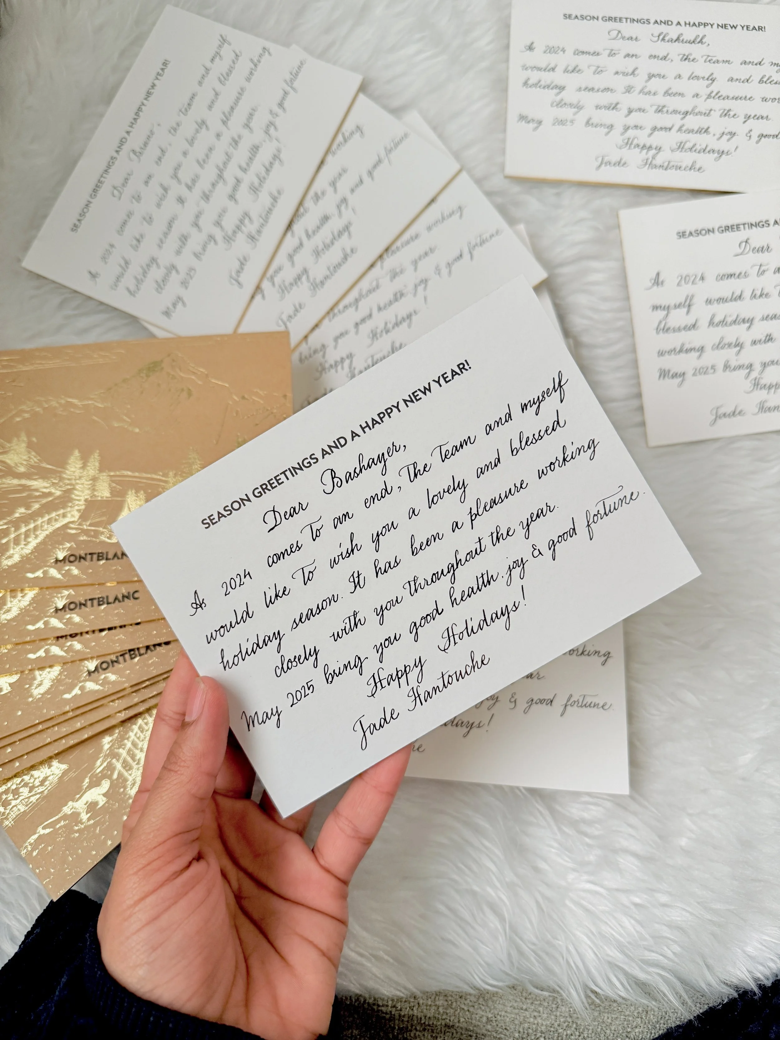 A person holding a white holiday greeting card with black cursive and bold print, surrounded by several similar cards and a gold embossed envelope, on a white furry surface.