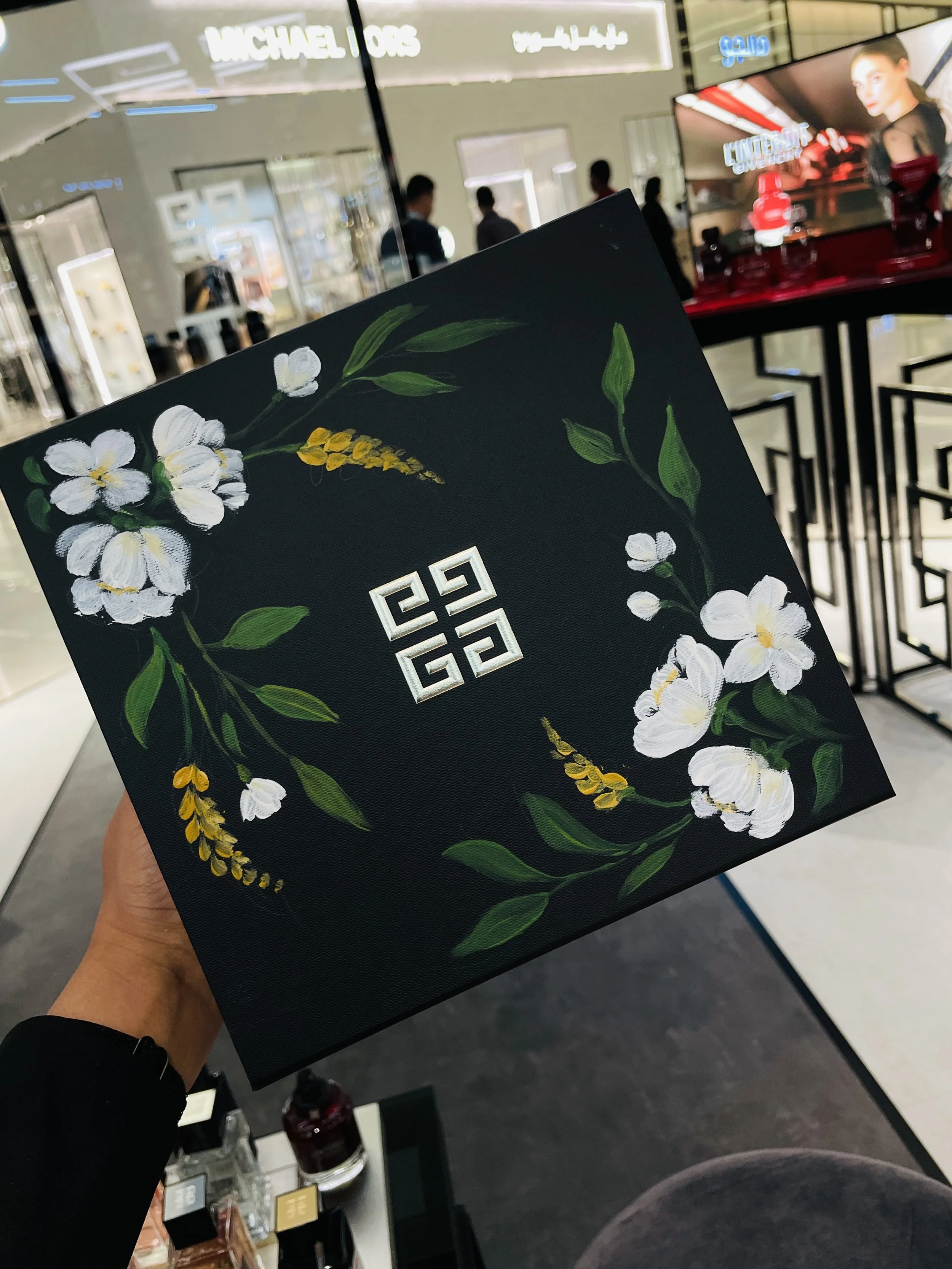 Close-up of a perfume box with floral artwork, being held in a shopping mall. The background shows a department store with people and digital screens.