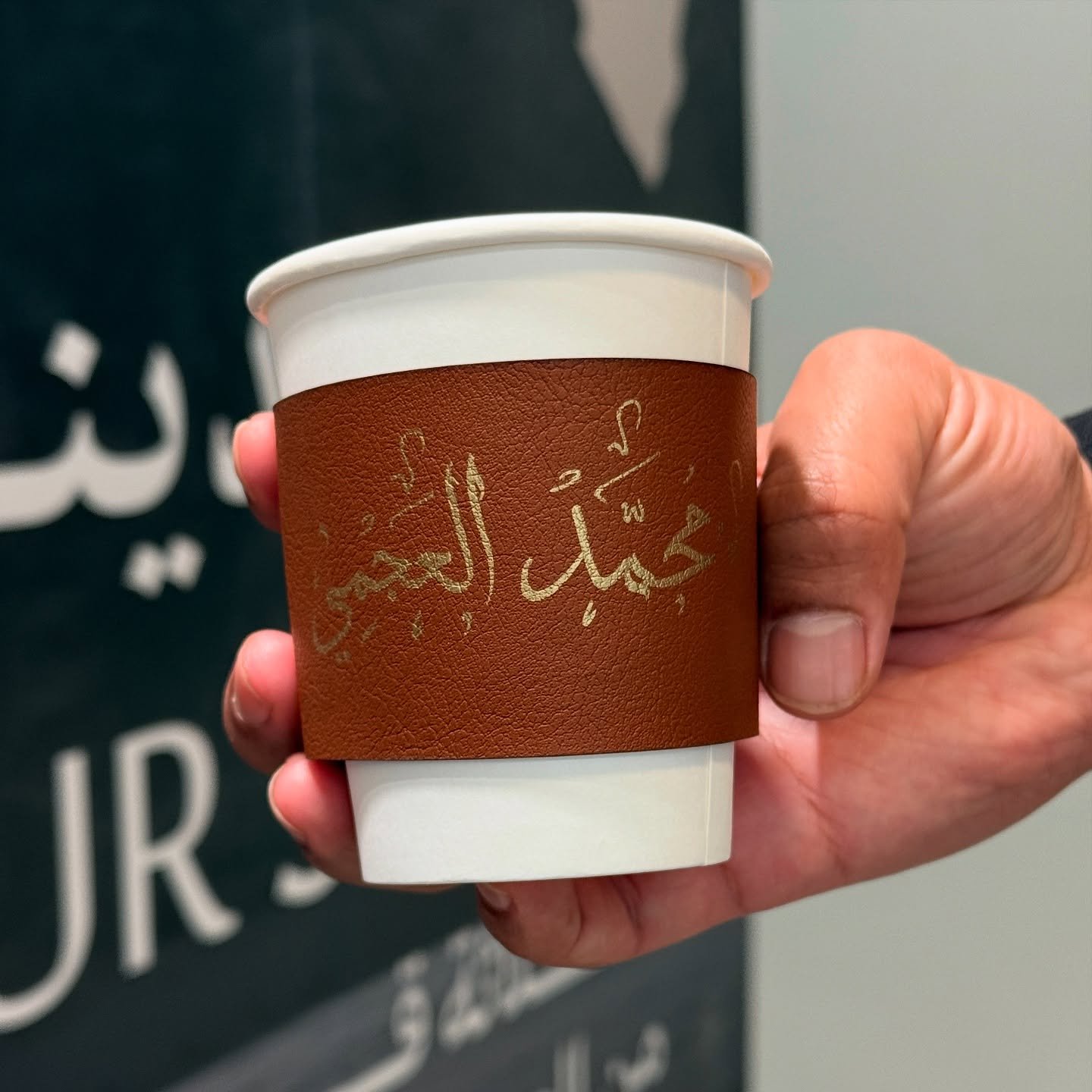 A person holding a disposable coffee cup with Arabic writing on a brown sleeve.