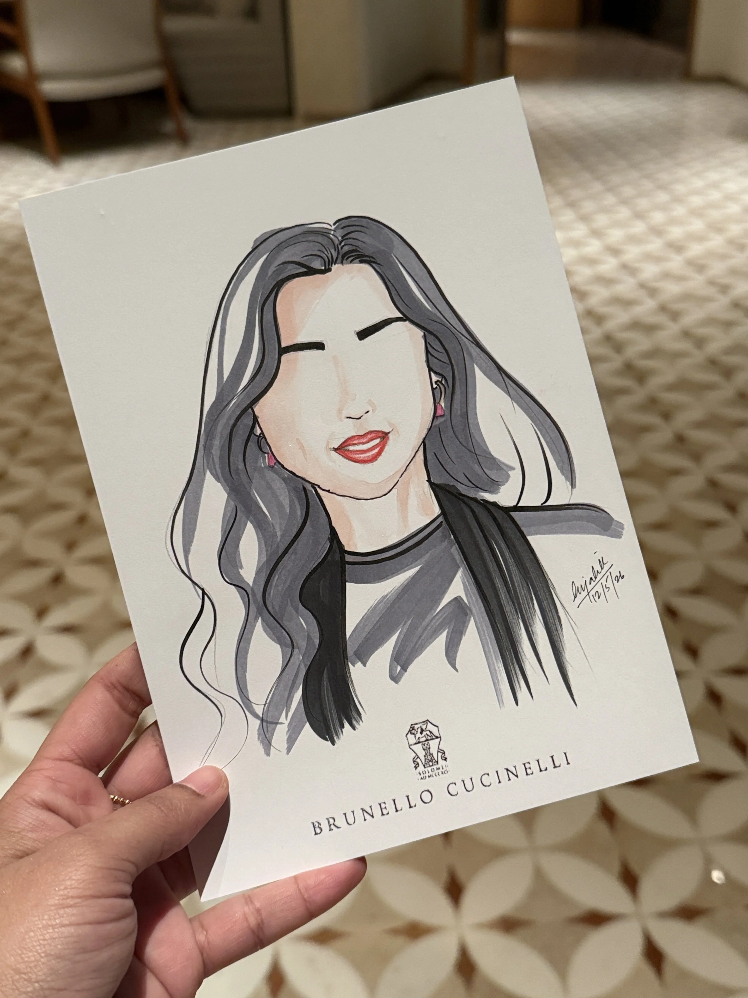 A person holding a printed booklet featuring a stylized illustration of a woman with long, flowing gray hair, closed eyes, and red lipstick. The booklet has the name 'Brunello Cucinelli' printed at the bottom, with logos and signatures above.