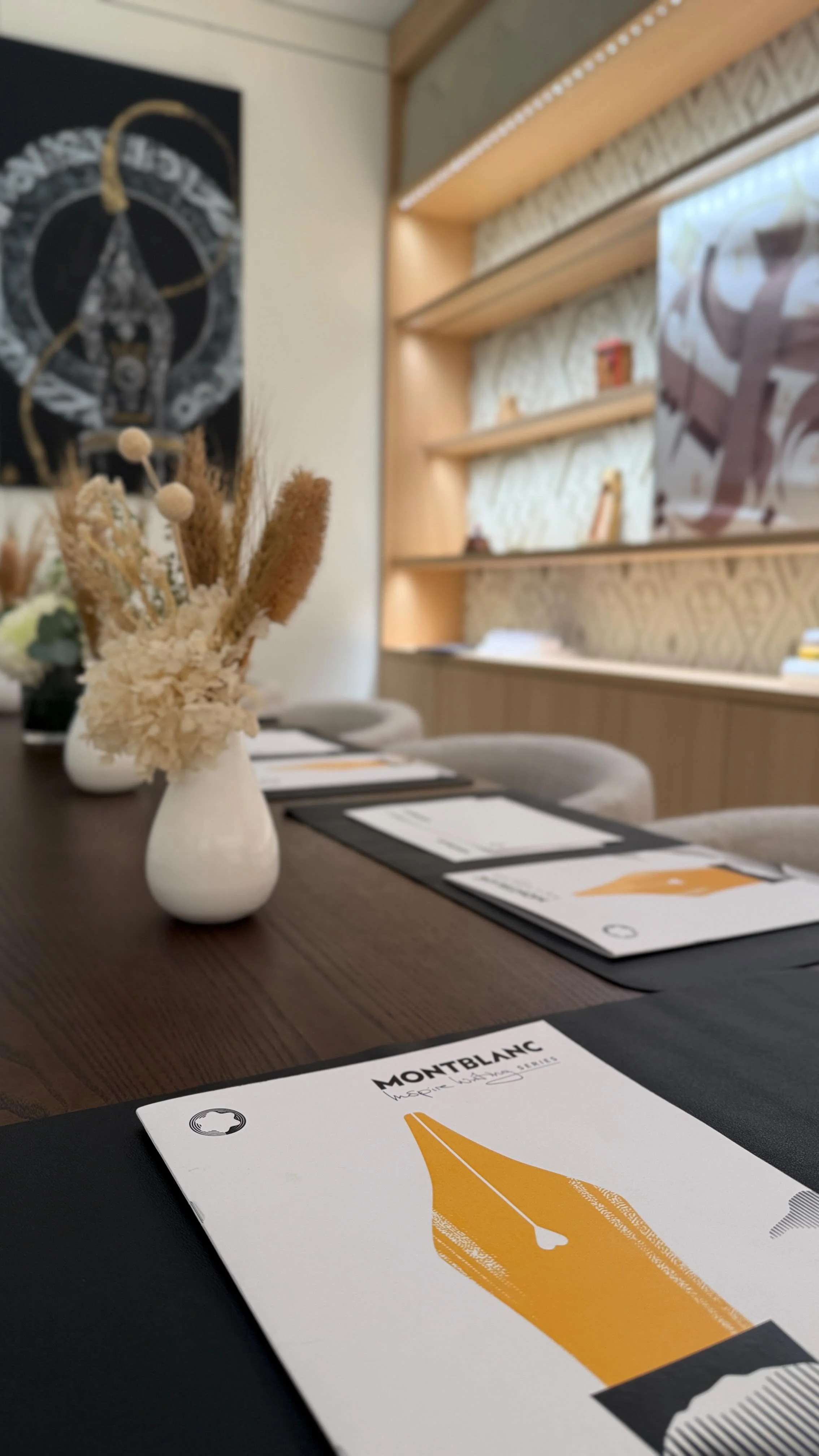 A table set with menus, a white ceramic vase with dried flowers, and seats in a modern dining or conference room. The room features a wooden shelving unit, decorative wall art, and a window with blinds.