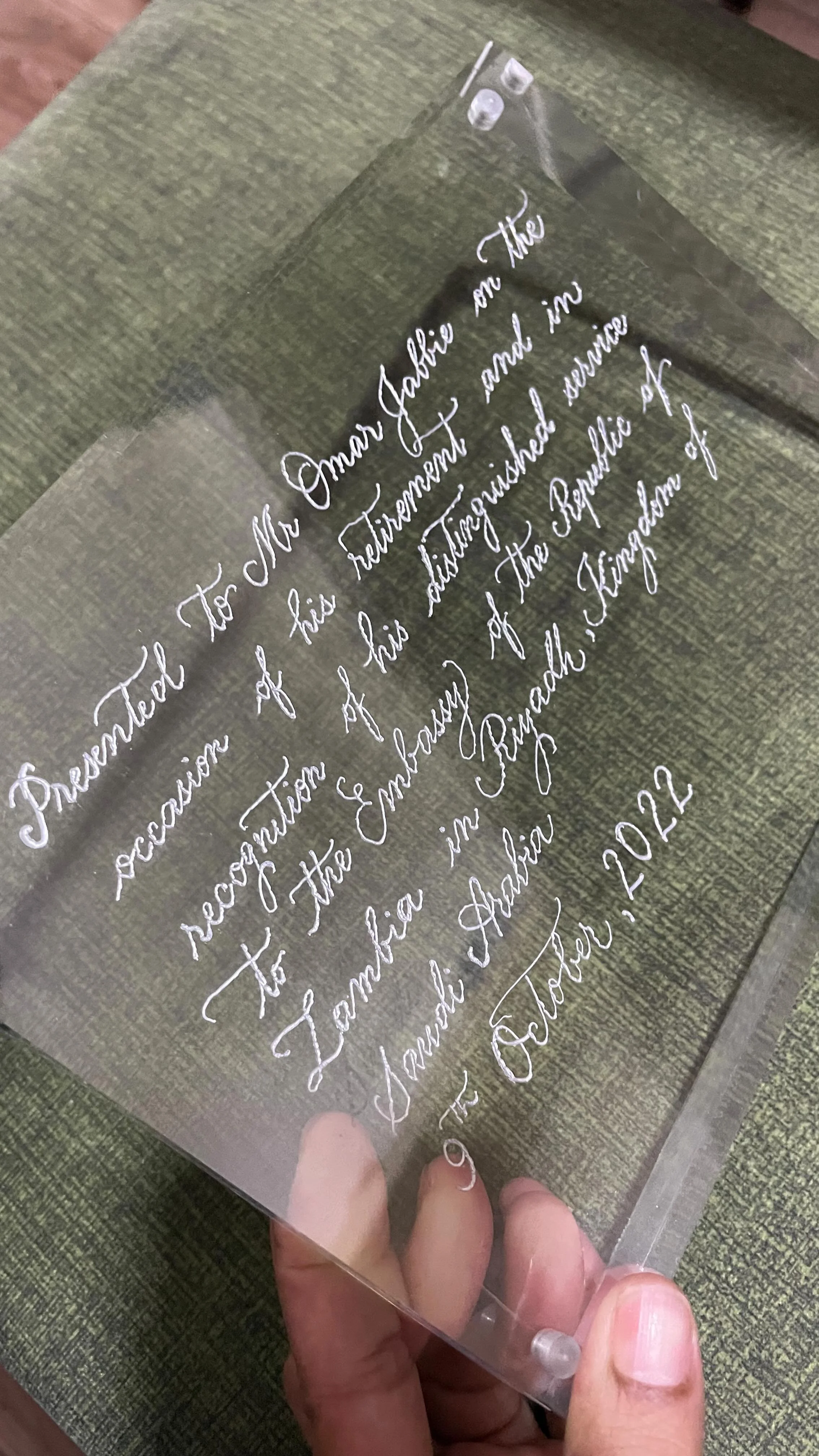 A person holding a transparent acrylic plaque with engraved cursive text on a green fabric surface.