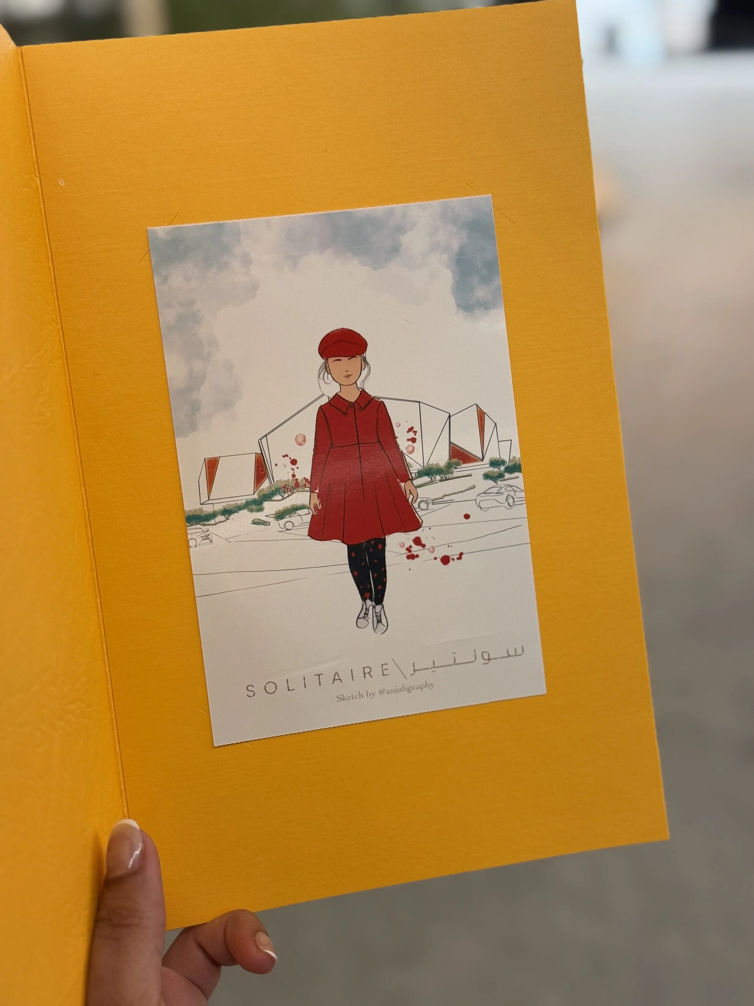 A person holding a yellow book or folder open to reveal a postcard or illustration of a woman in red clothing and a red beret, standing in front of modern architectural structures with clouds in the sky above. The text on the card reads 'SOLITAIRE' a