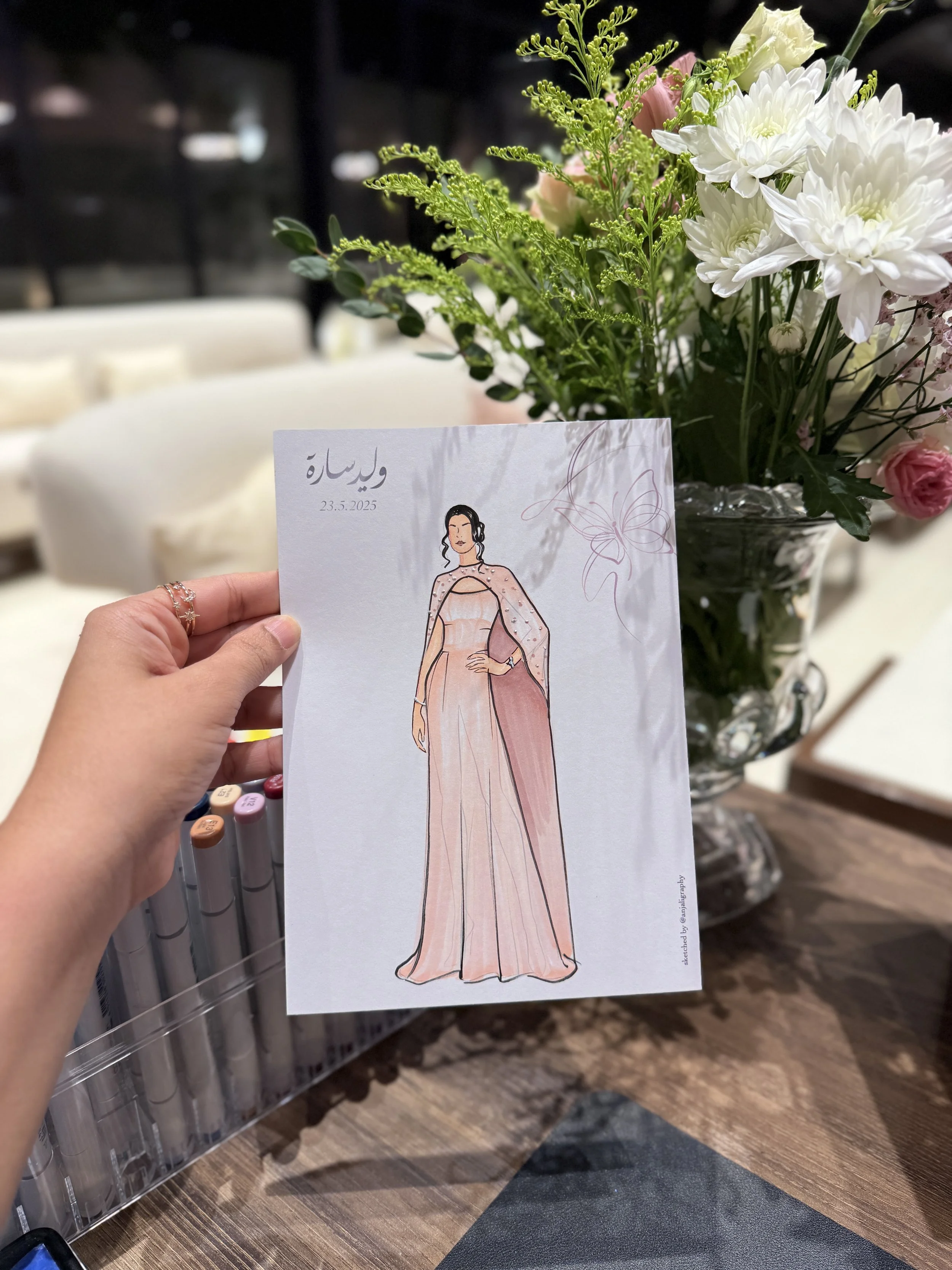 A hand holding a sketch of a woman in a pink evening gown and cape in front of a flower arrangement with white, pink, and yellow flowers.
