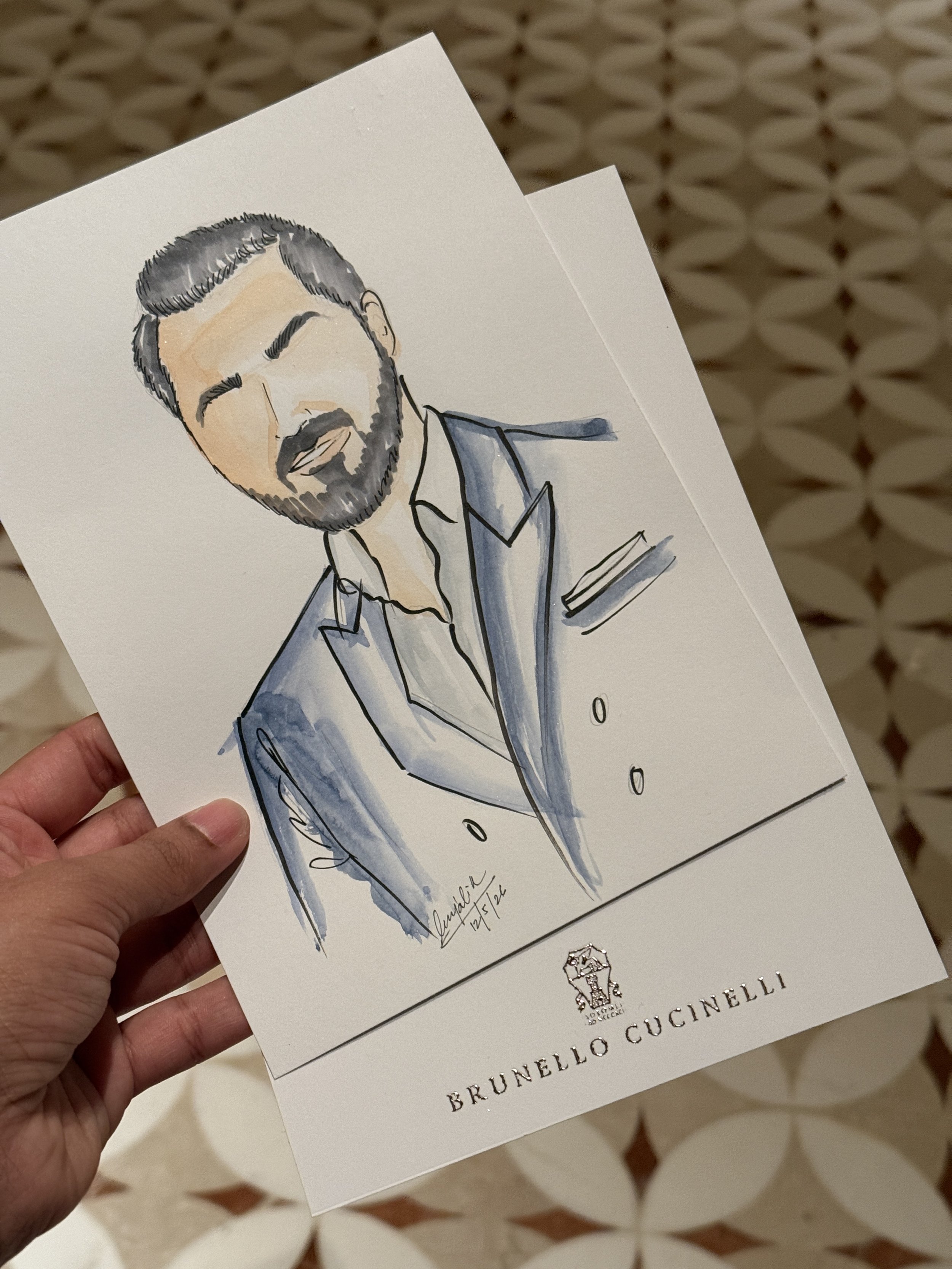Watercolor portrait of a man with dark hair, a beard, and closed eyes on a card held in hand, with the text 'Brunello Cucinelli' printed at the bottom.
