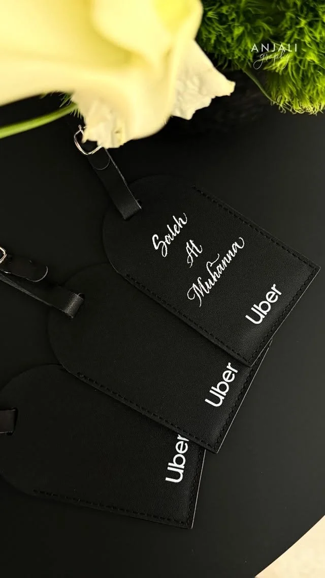 Three black Uber tags labeled 'Saudha Al Muhanna' on a black surface, with white flowers and green foliage in the background.