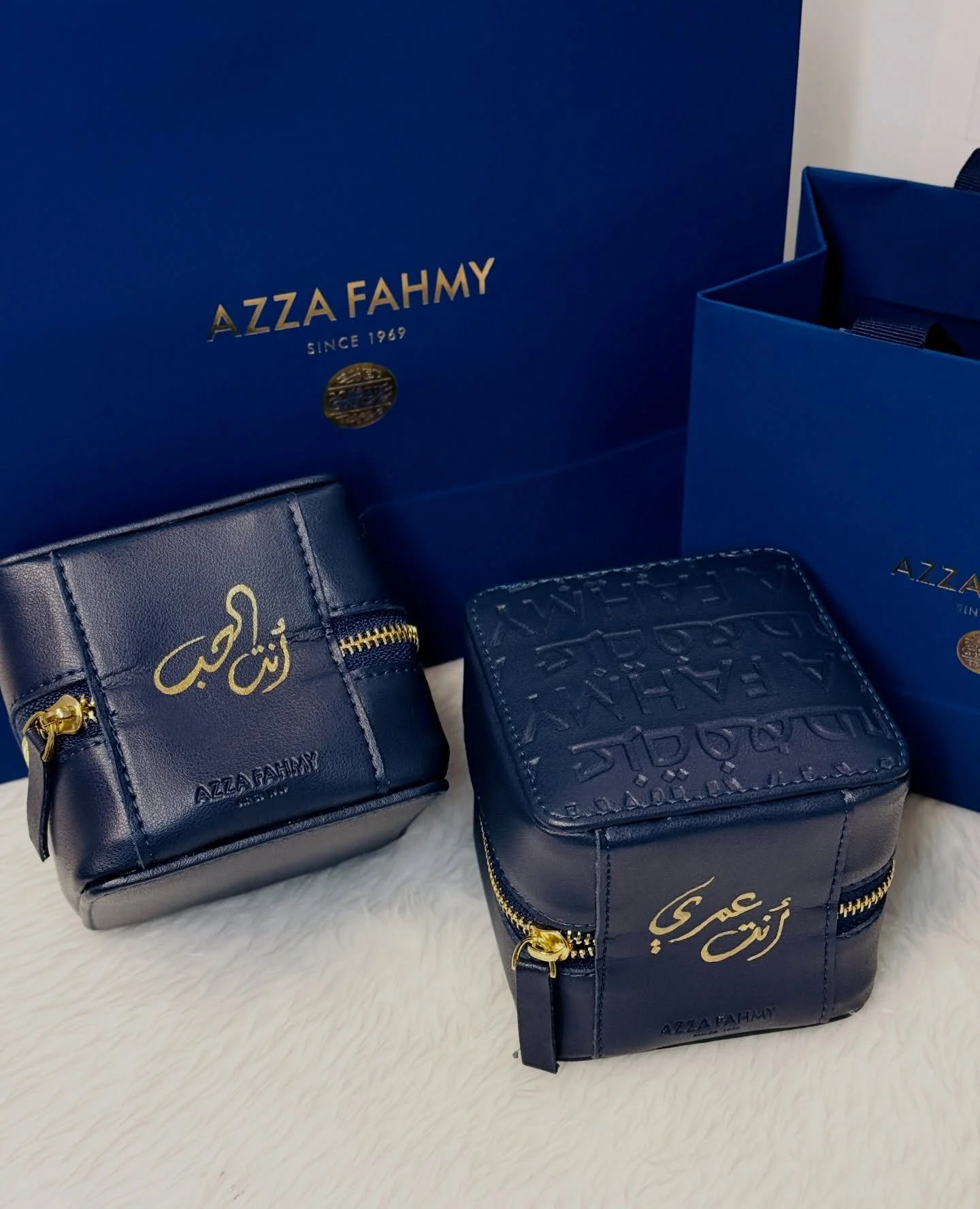 Two navy blue handbags with gold zippers and Arabic calligraphy, placed on a white furry surface. Behind them are two large blue boxes with gold lettering that reads "AZZA FAHMY SINCE 1969" and a small gold emblem.