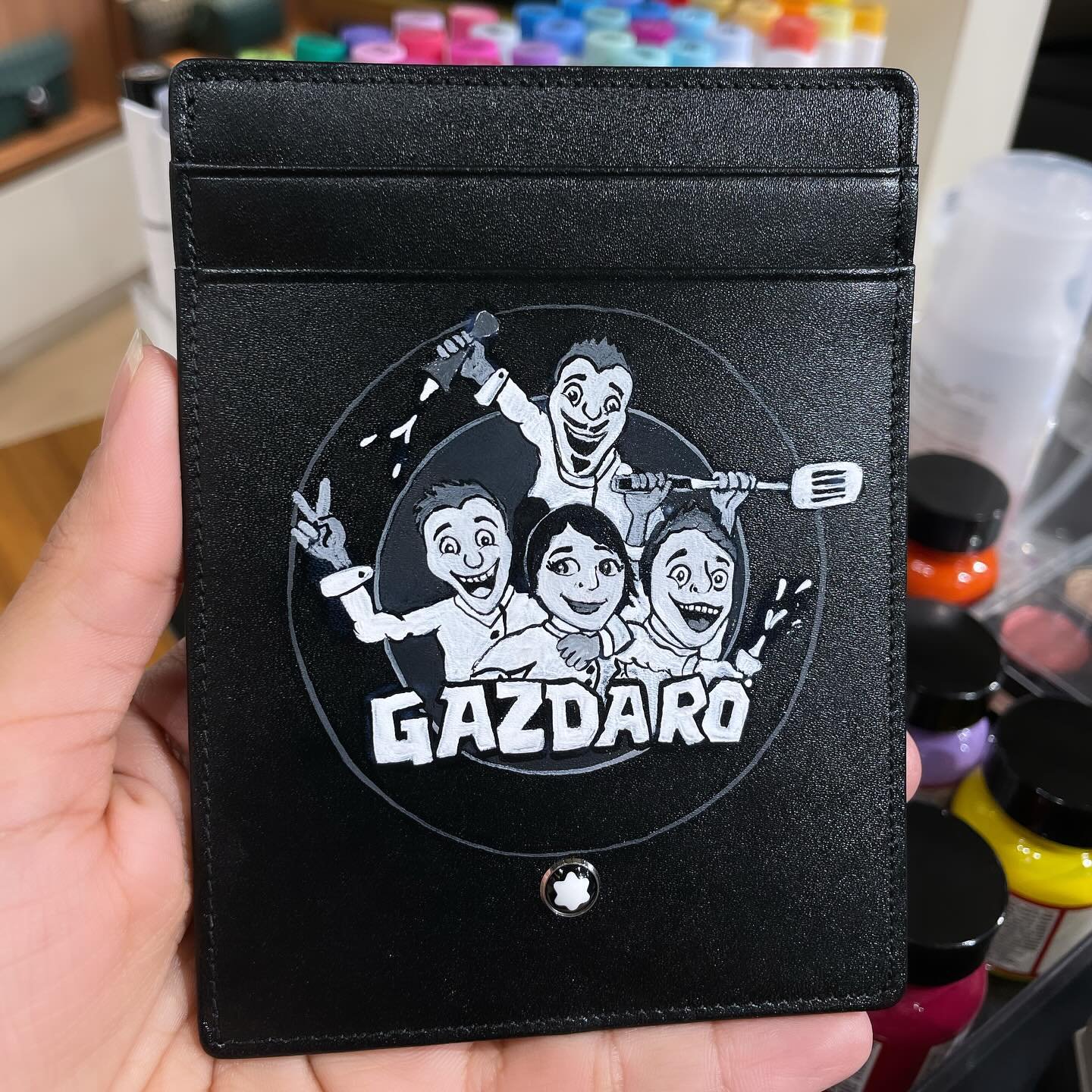 A black wallet featuring a cartoon illustration of four cheerful people holding tools, with the word "GAZARO" underneath.