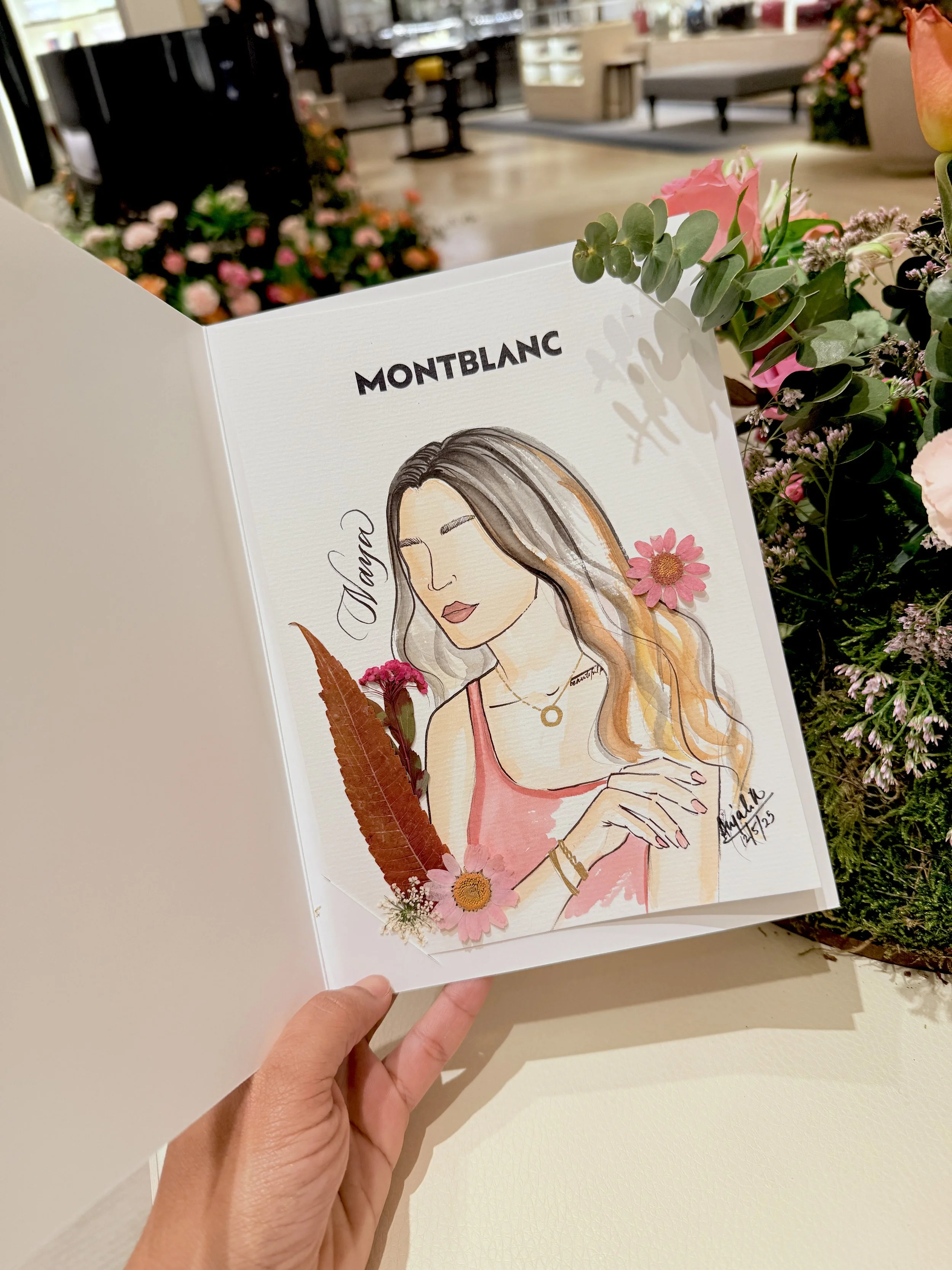 A hand holding a sketchbook with a colorful illustrated portrait of a woman, surrounded by pink flowers and green leaves, in a store or cafe setting.