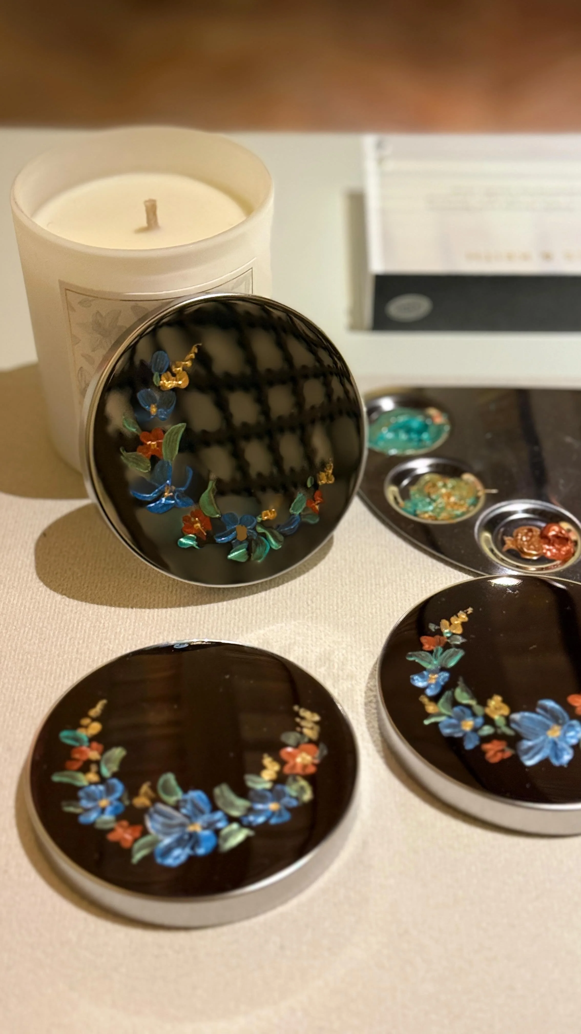 Collection of jewelry including floral earrings with blue, orange, green, and gold accents, displayed on black round and oval surfaces, with a white candle in the background.