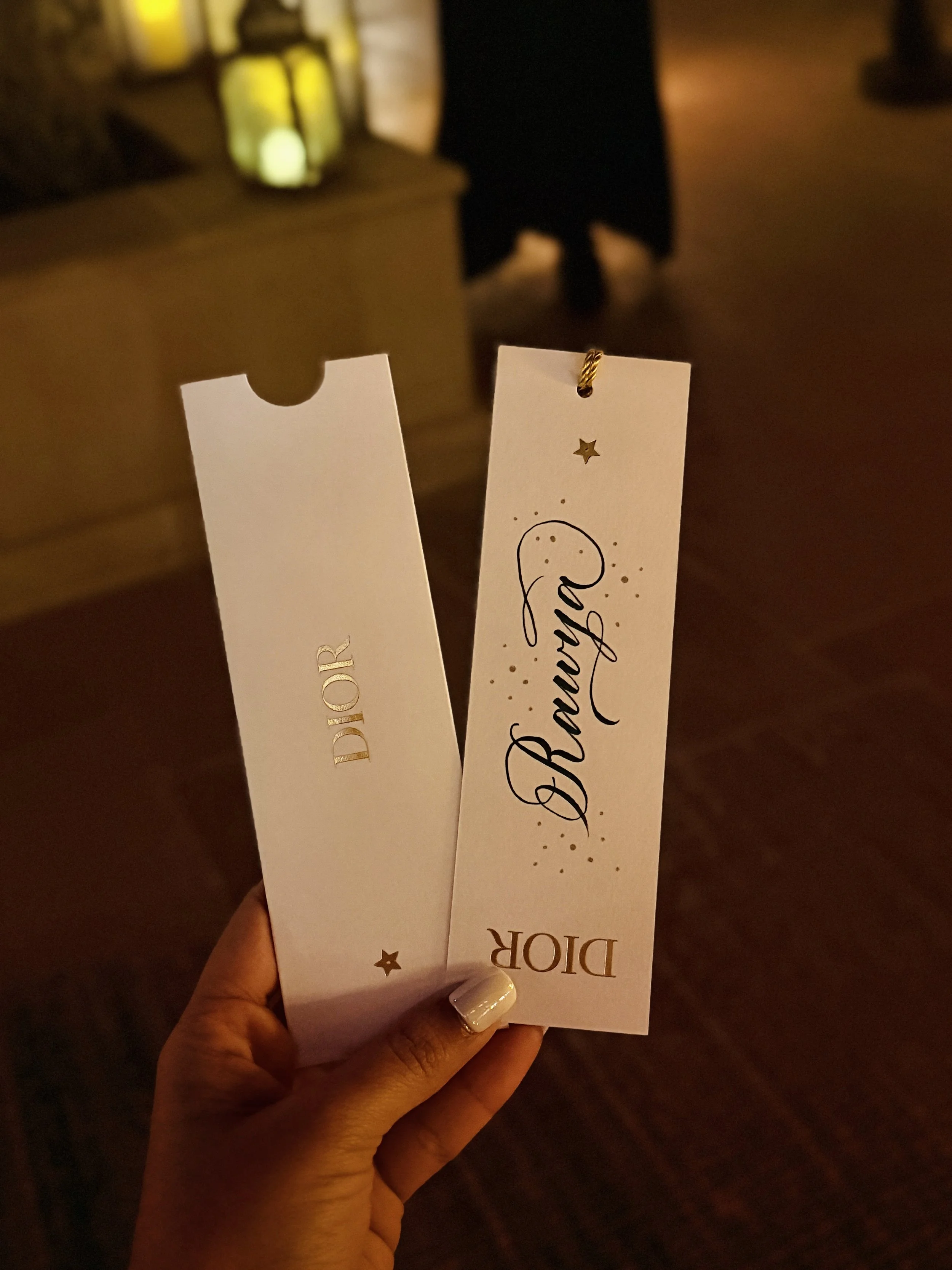 Person holding two white event tickets with 'Dior' written on them, in a dimly lit room.
