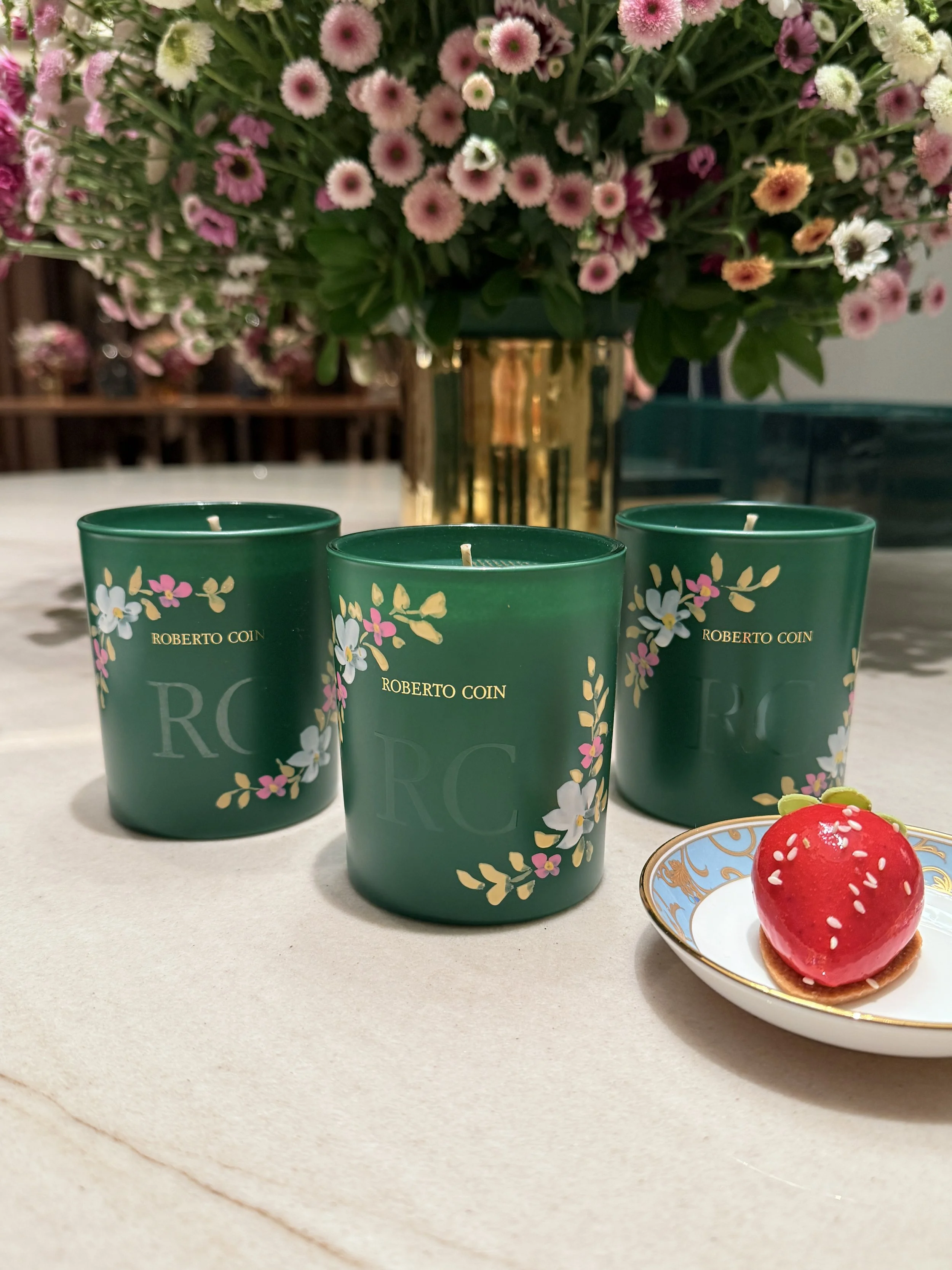 Three green scented candles with floral designs in the foreground, a gold vase of pink and white flowers in the background, and a small red dessert with white sprinkles on a decorative plate to the right.