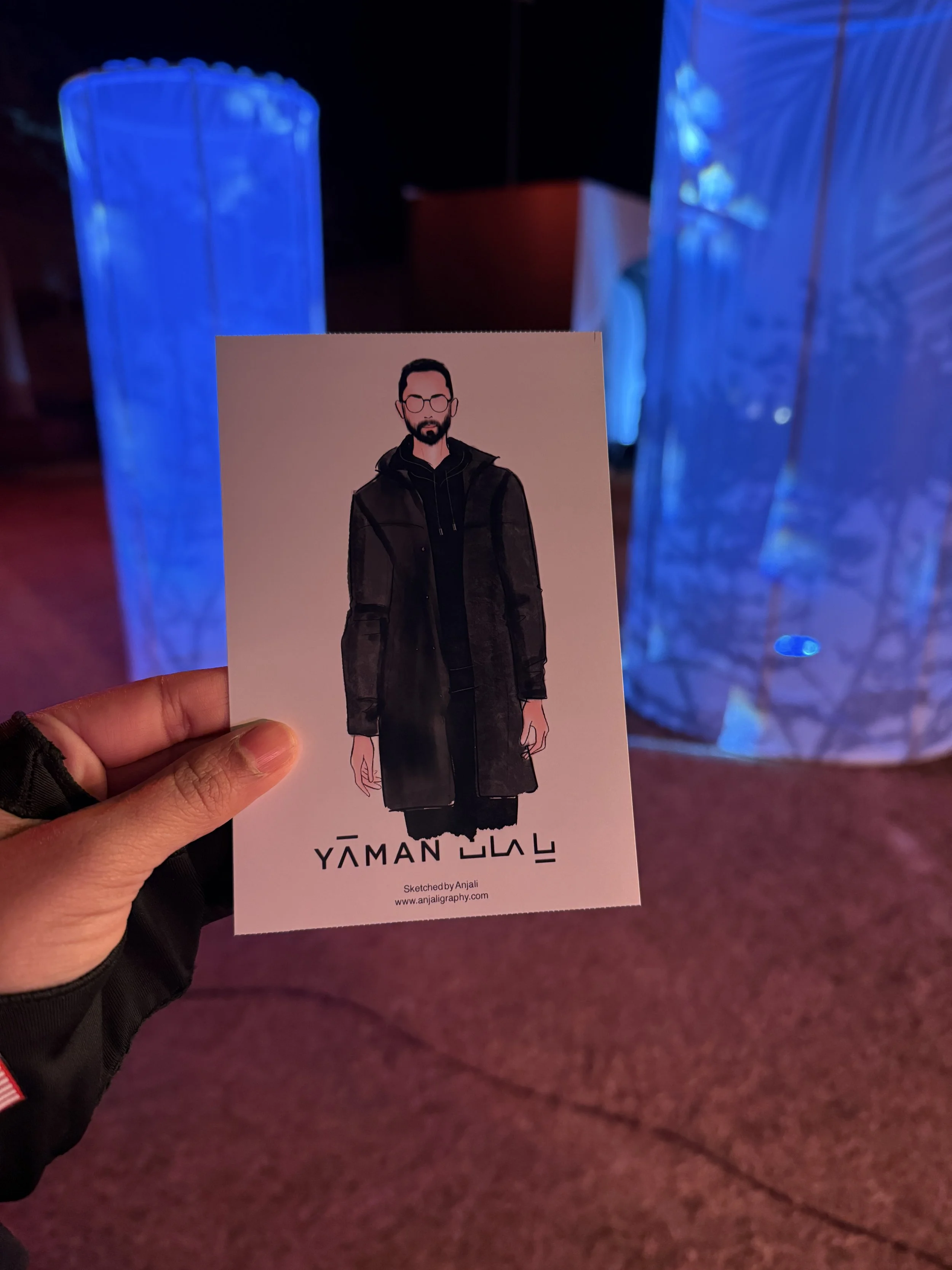 A hand holding a graphic design card with an illustrated man wearing glasses, a black jacket, and a black hoodie, against a background of blue illuminated objects in a dark space.