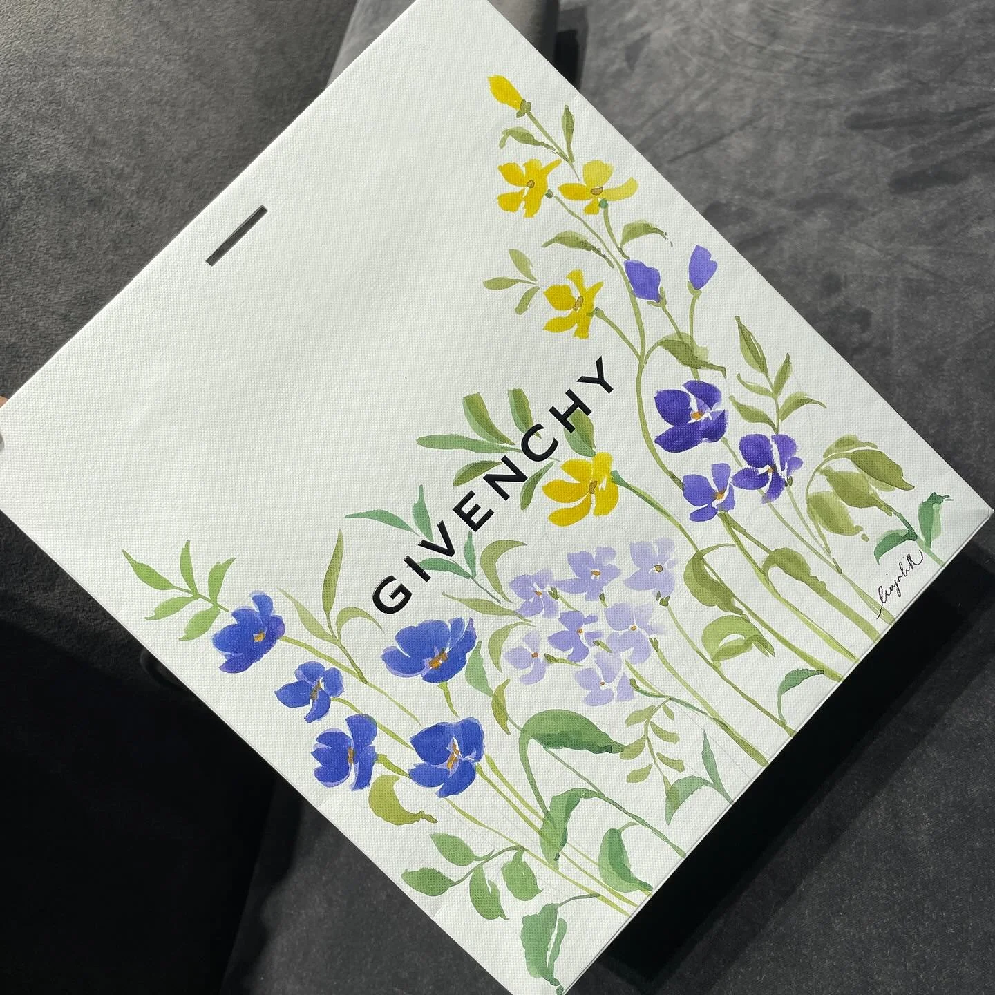 A white card with yellow and purple painted flowers and green leaves, the word "GIVENCHY" in black on the front, and a signature in the bottom right corner.