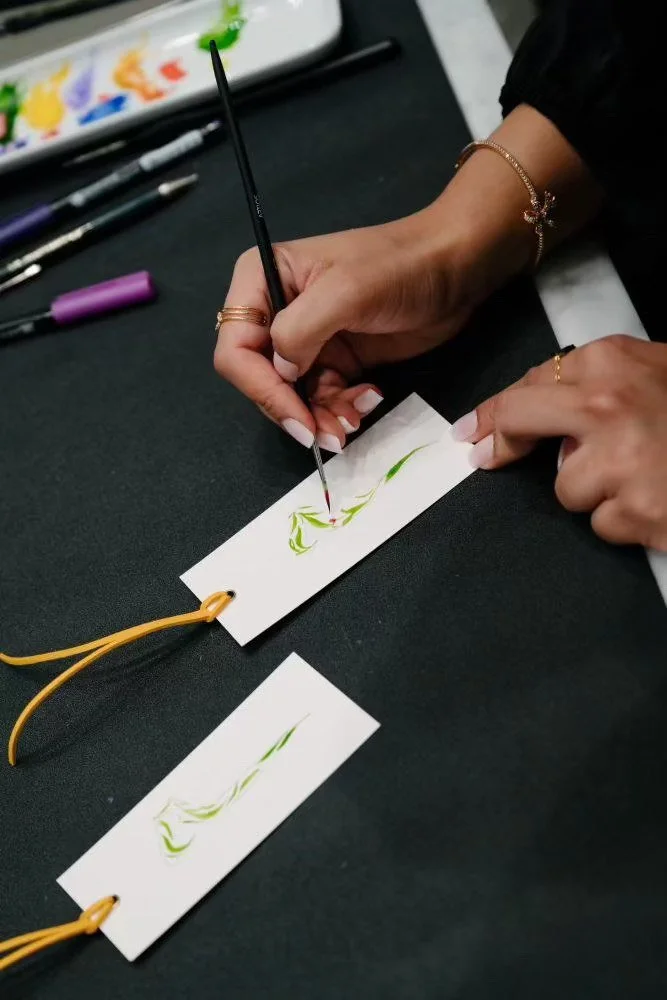 Person decorating paper tags with green and red paint, using a fine brush, on a black surface with art supplies visible.