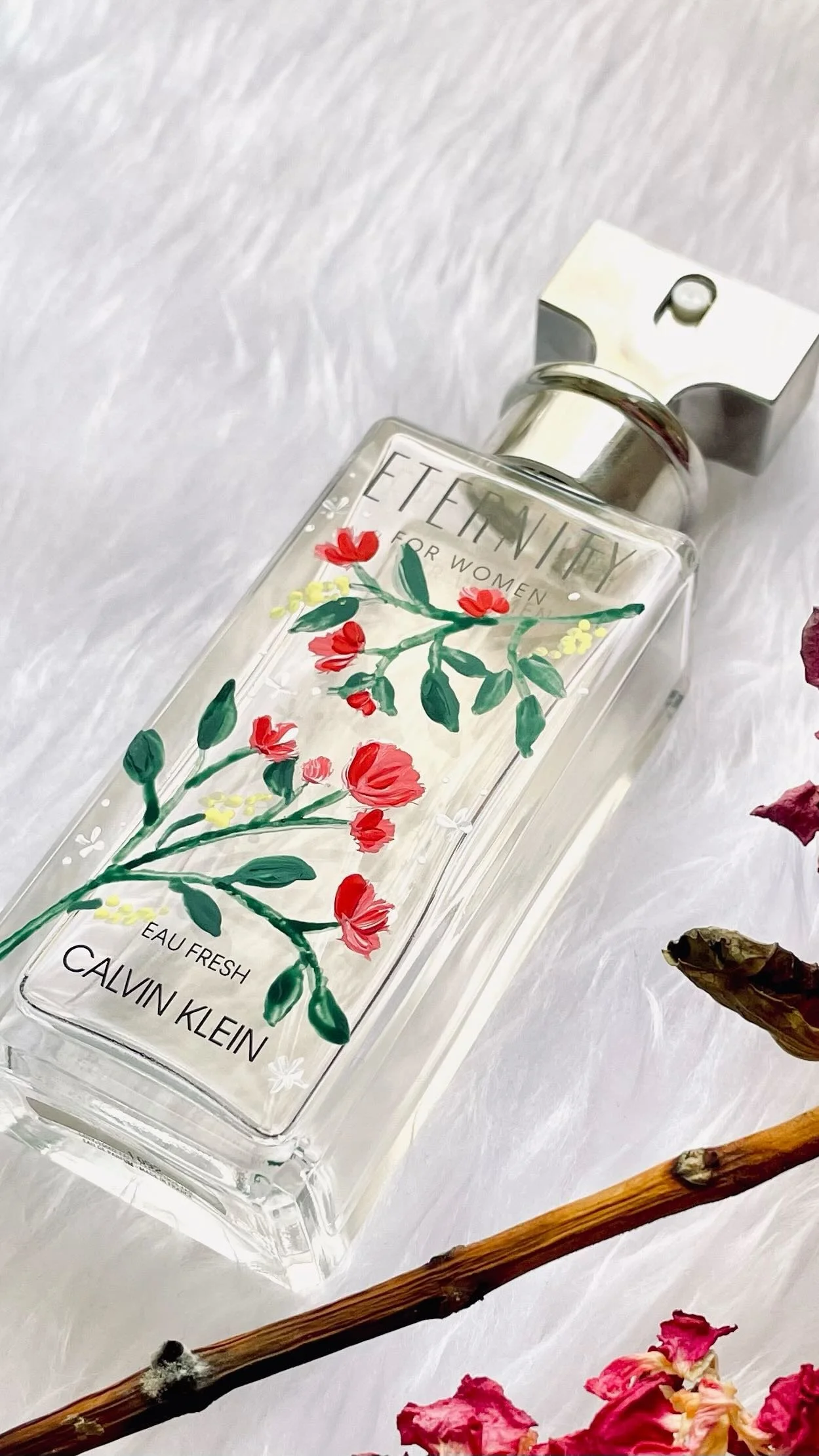 A Calvin Klein perfume bottle called 'Eternity for Women' with floral artwork on its front, placed on a white textured surface, surrounded by pink and purple dried flowers and a small branch.