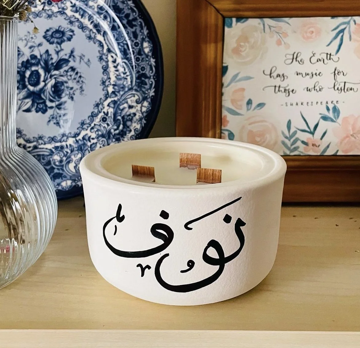 A white candle holder with Arabic calligraphy on it, placed on a wooden surface. In the background, there are decorative items including a framed quote that says, "The Earth has music for those who listen" by Shakespeare, a blue and white patterned p