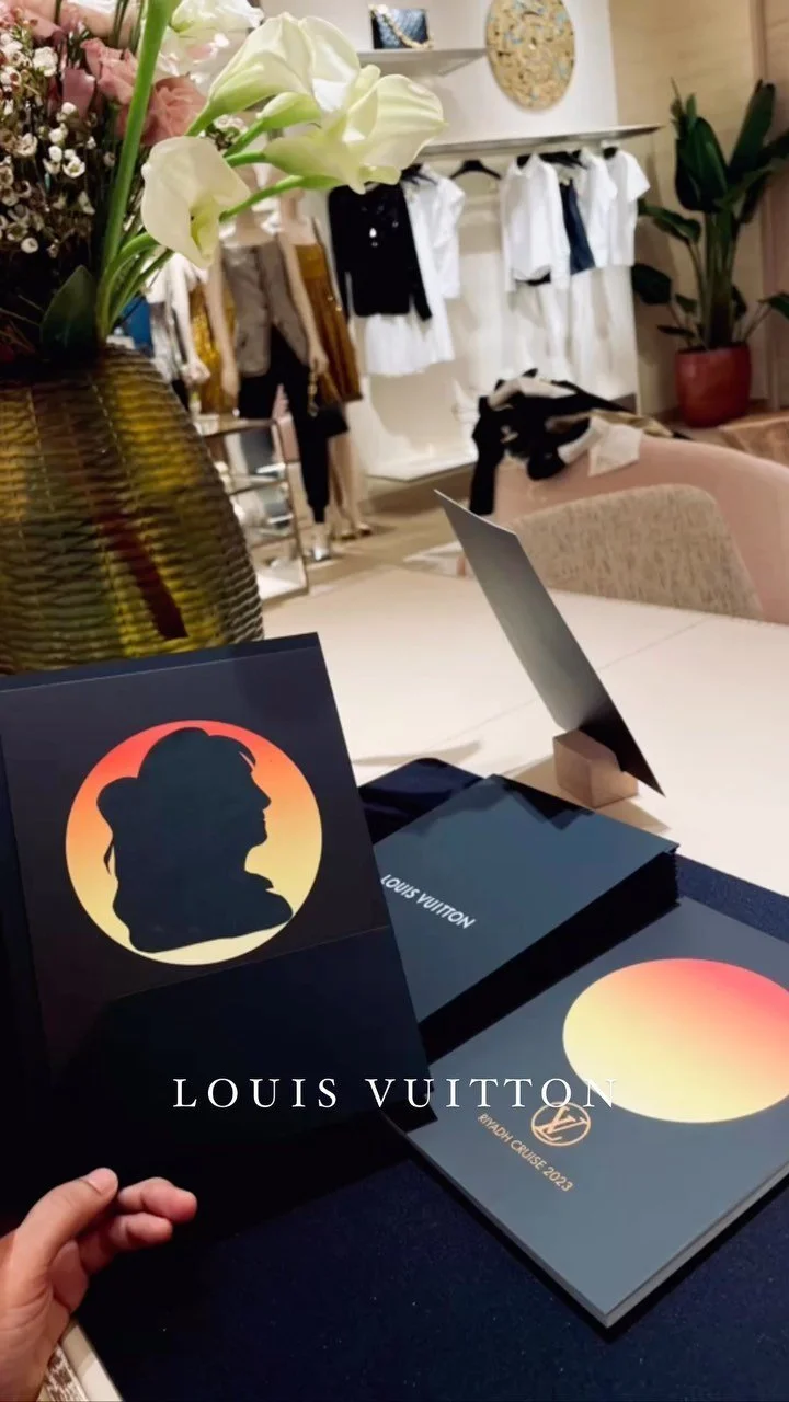 A table with Louis Vuitton brochures, a woman’s silhouette logo display, a small photo frame, and a floral arrangement in a vase. In the background, clothing is hanging in a boutique or showroom.