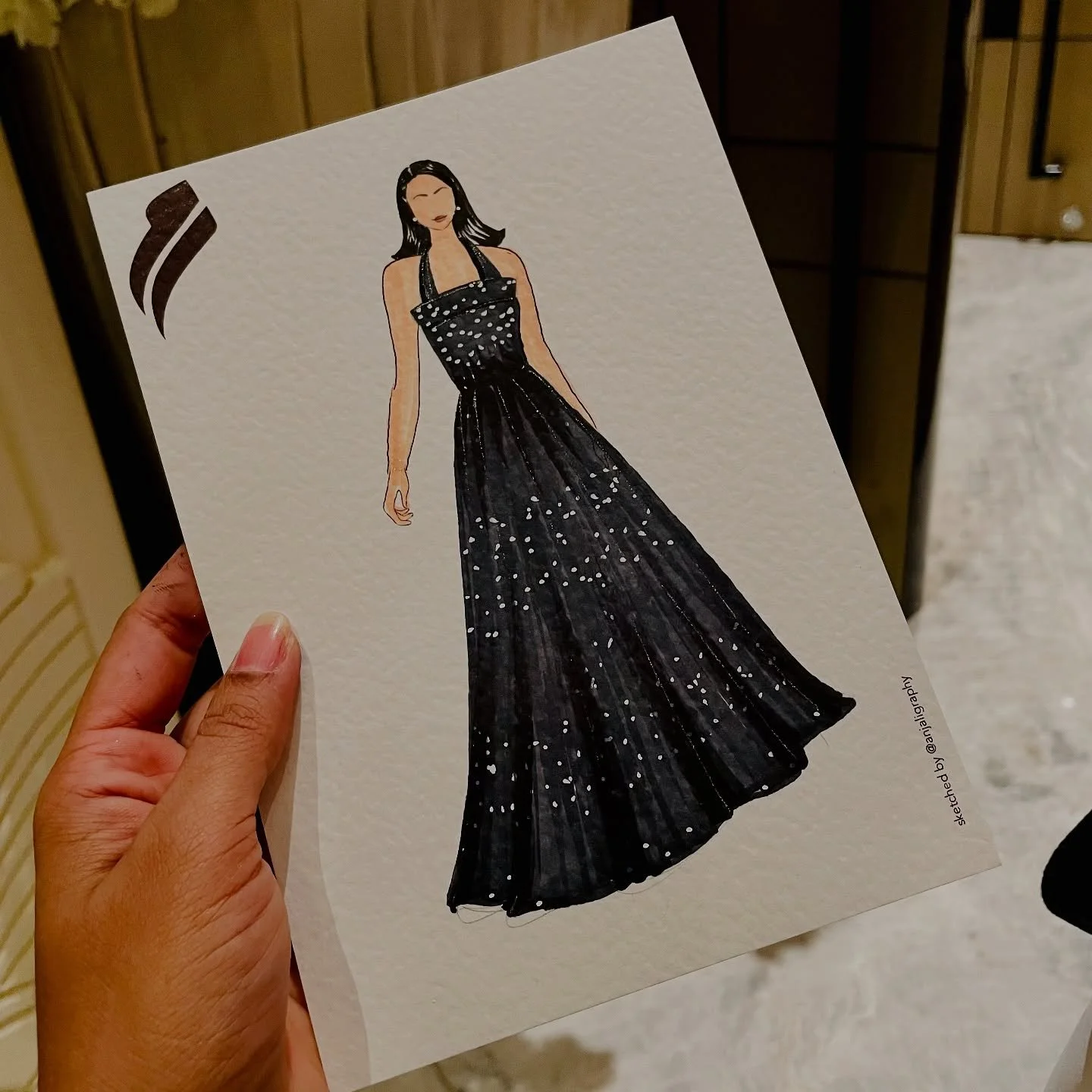 Hand holding a fashion illustration of a woman in a black, sparkling evening gown with a halter neckline and long flowing skirt.