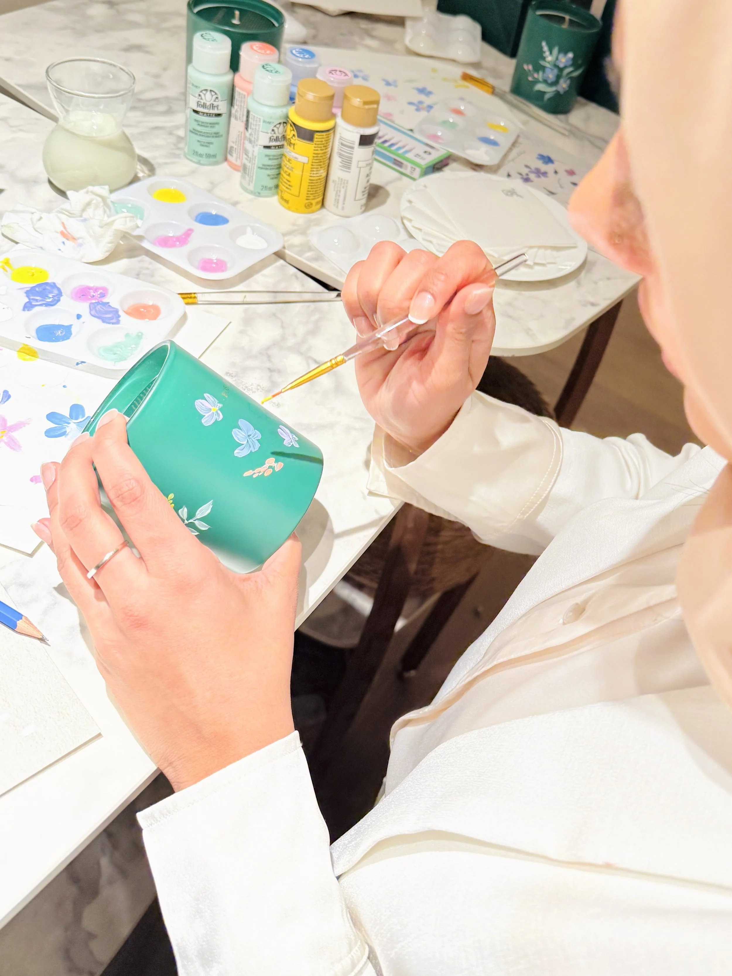 A person painting a ceramic mug with floral decorations using a small brush, surrounded by various paint bottles, palettes, and painting supplies on a table.