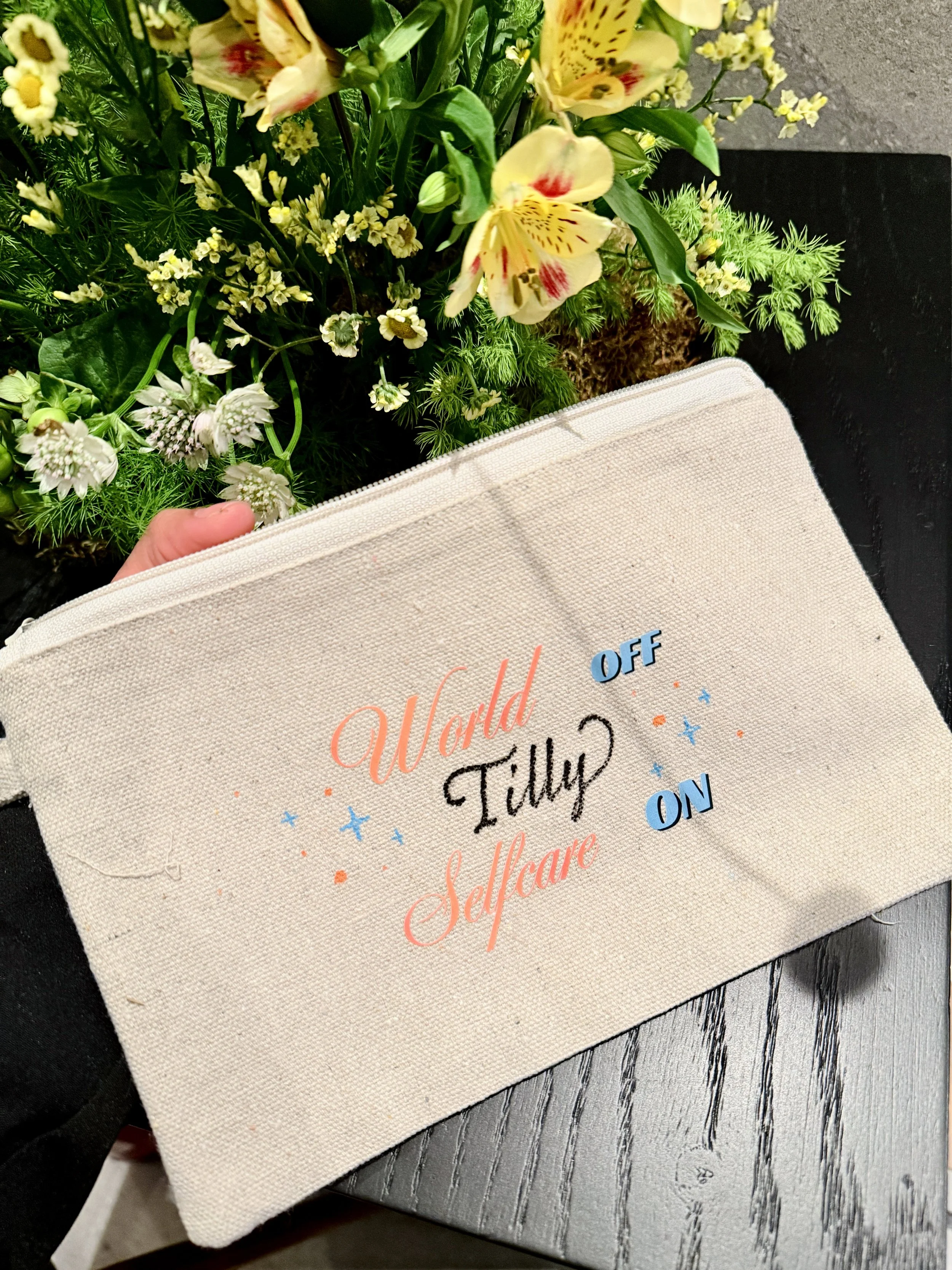 Close-up of a floral arrangement with yellow and white flowers, placed on a gray wooden surface, partially covered by a beige fabric tote with colorful text.