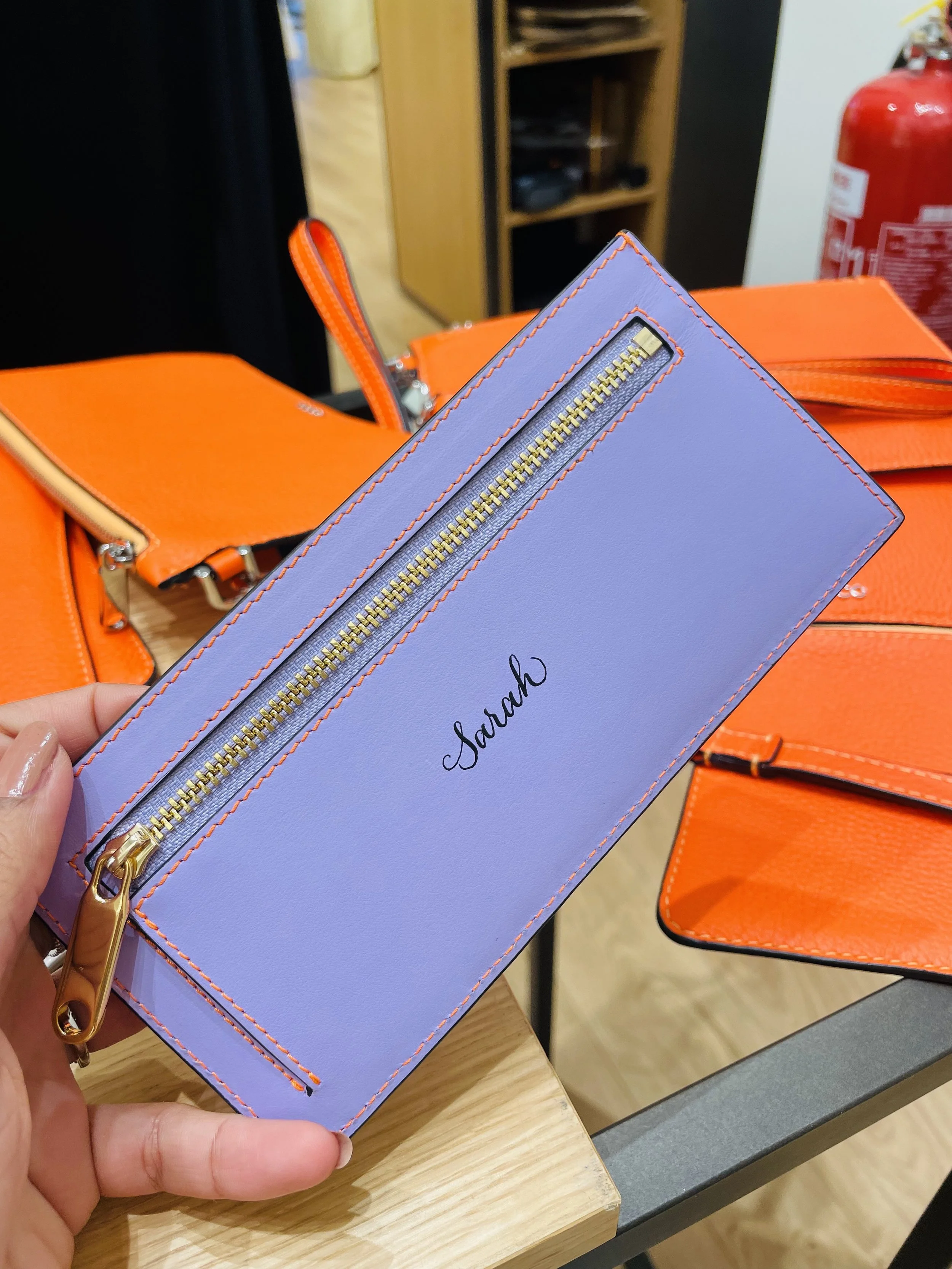 A lavender-colored leather pouch with a gold zipper and the name 'Sarah' written in black, held by a person's hand, with orange handbags and a fire extinguisher in the background.