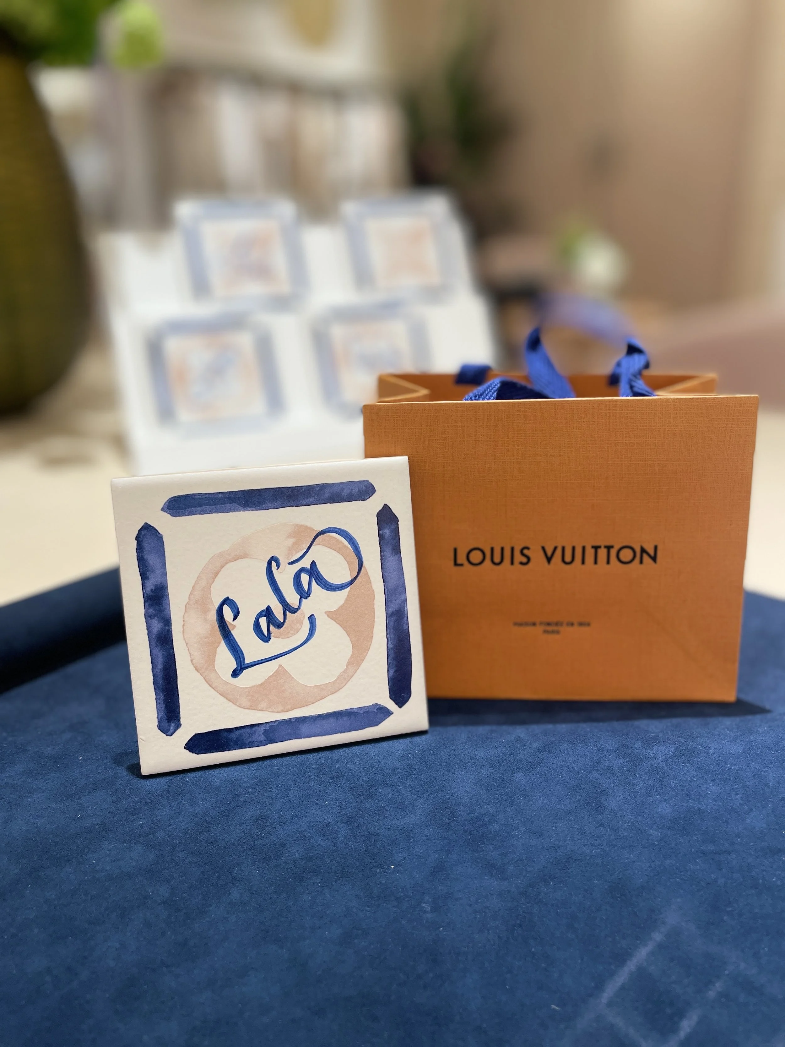 A small white box with blue handwritten logo saying 'Lala' next to an orange Louis Vuitton shopping bag with blue ribbons on a dark surface.