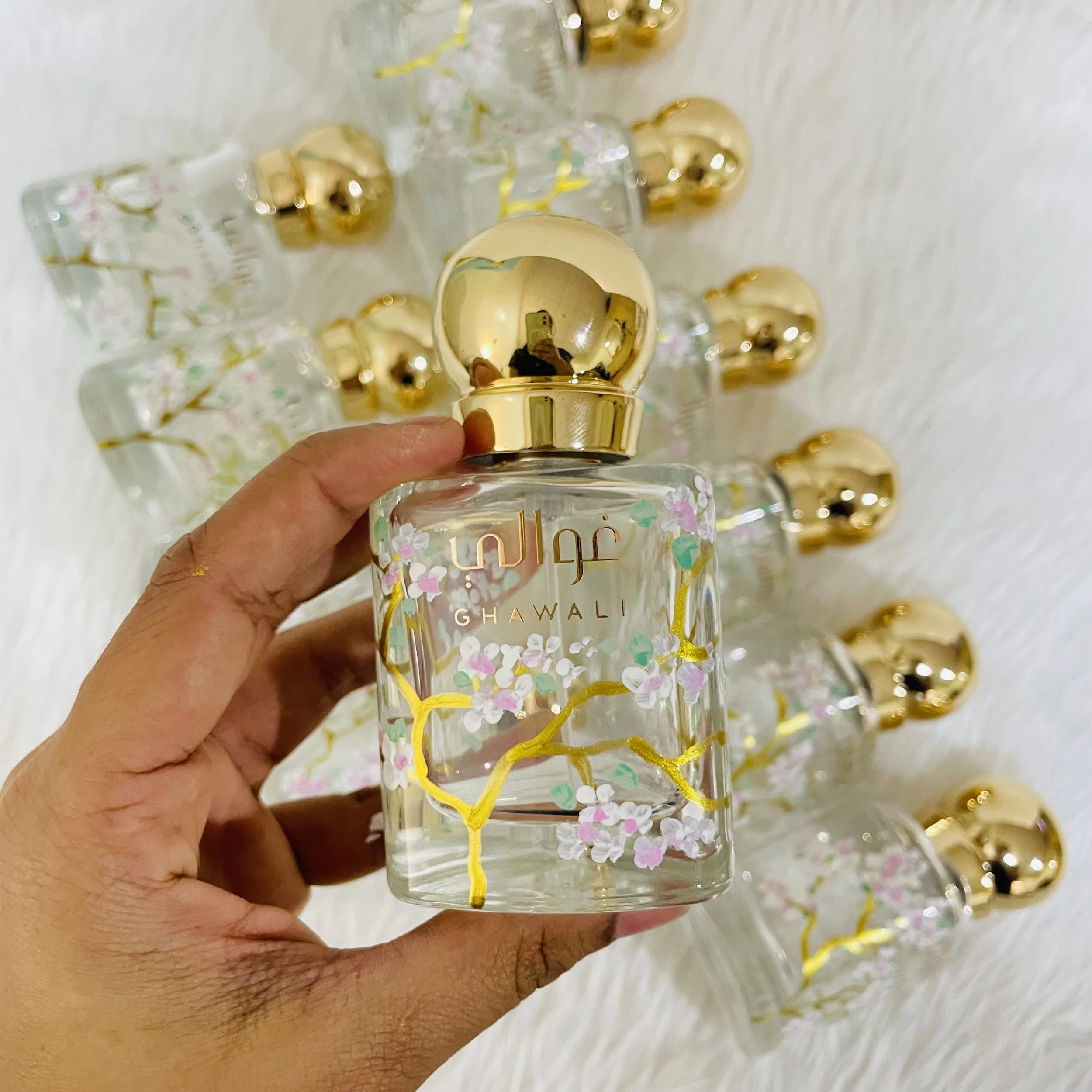 A person holding a clear glass bottle with a gold-colored cap, decorated with pink, green, and white floral patterns. The background shows multiple similar bottles.