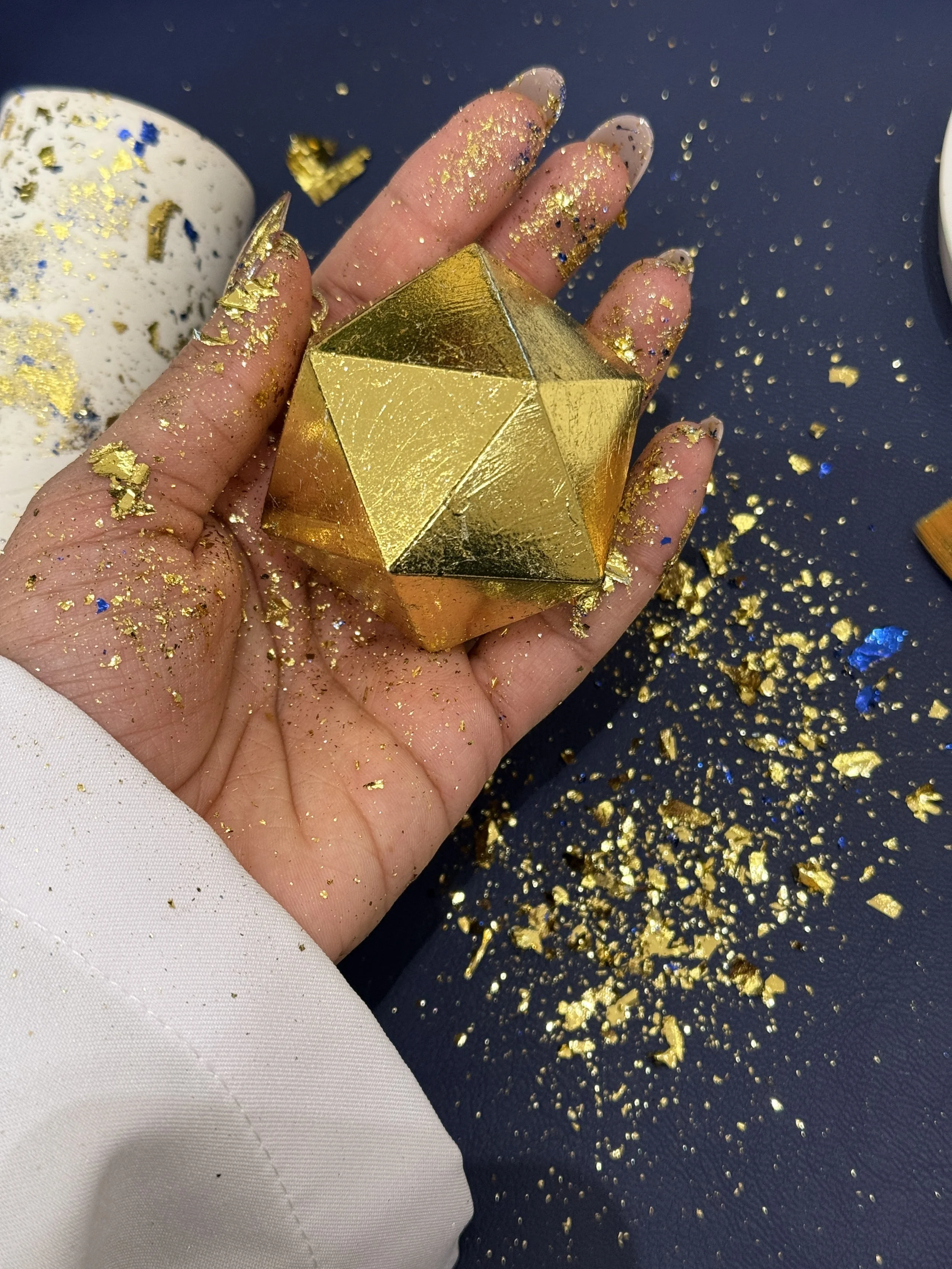 A hand holds a shiny gold geometric object, with gold foil and glitter on fingers and surrounding surface.