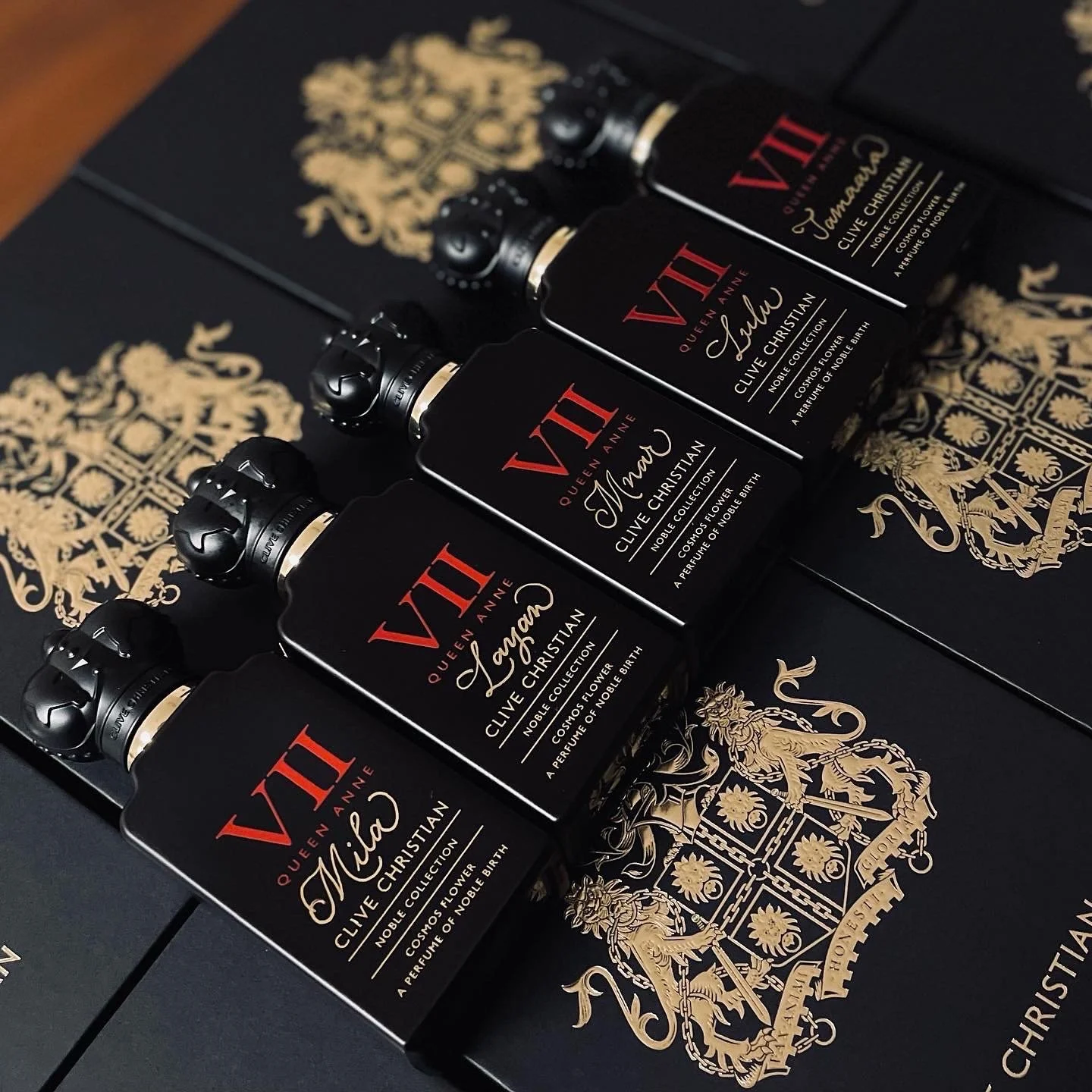 Four black perfume bottles with gold and red labels, positioned in a diagonal row on black boxes with gold crest designs.